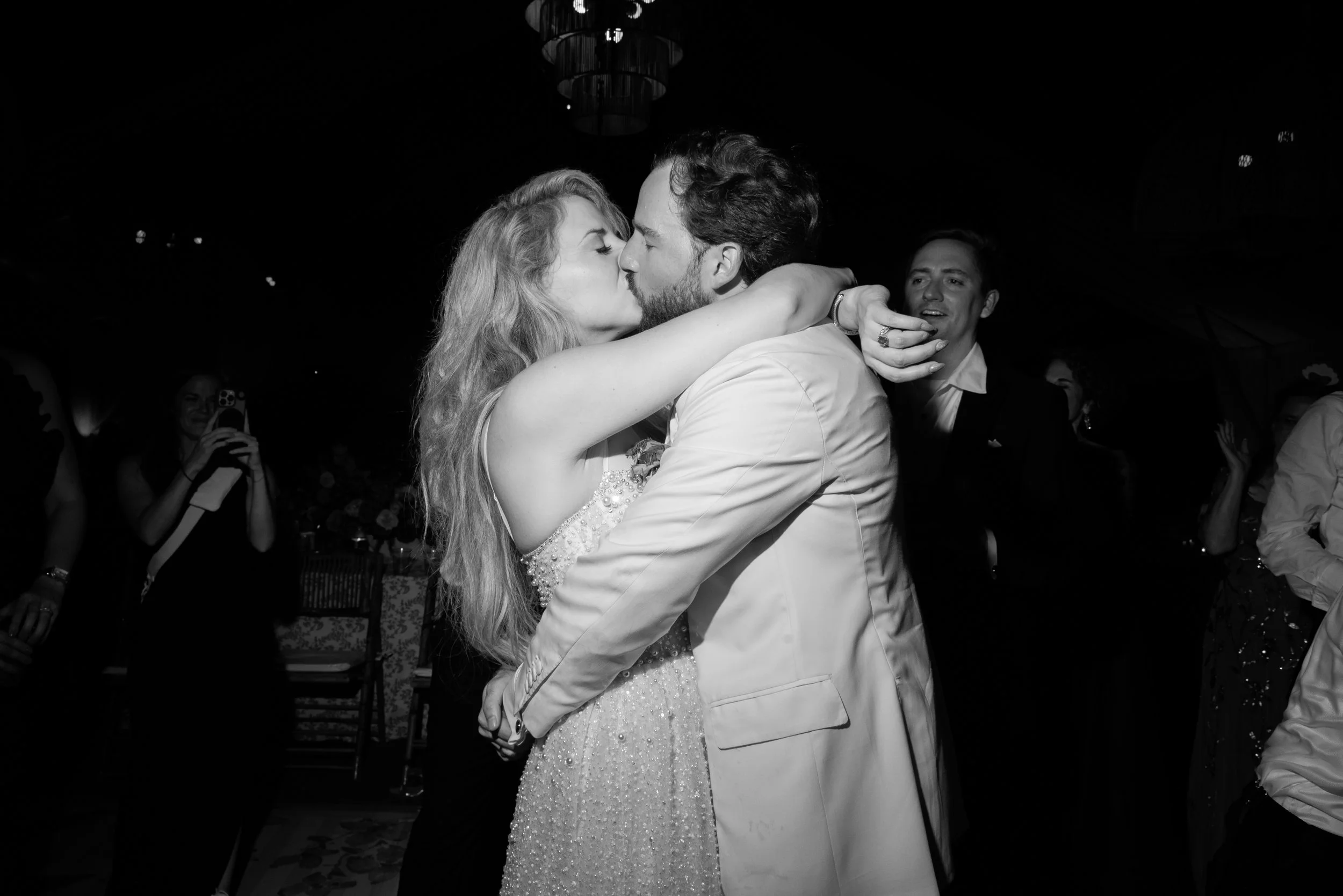 Black-and-white photograph of Katie and Trevor kissing on the dance floor as guests celebrate around them.