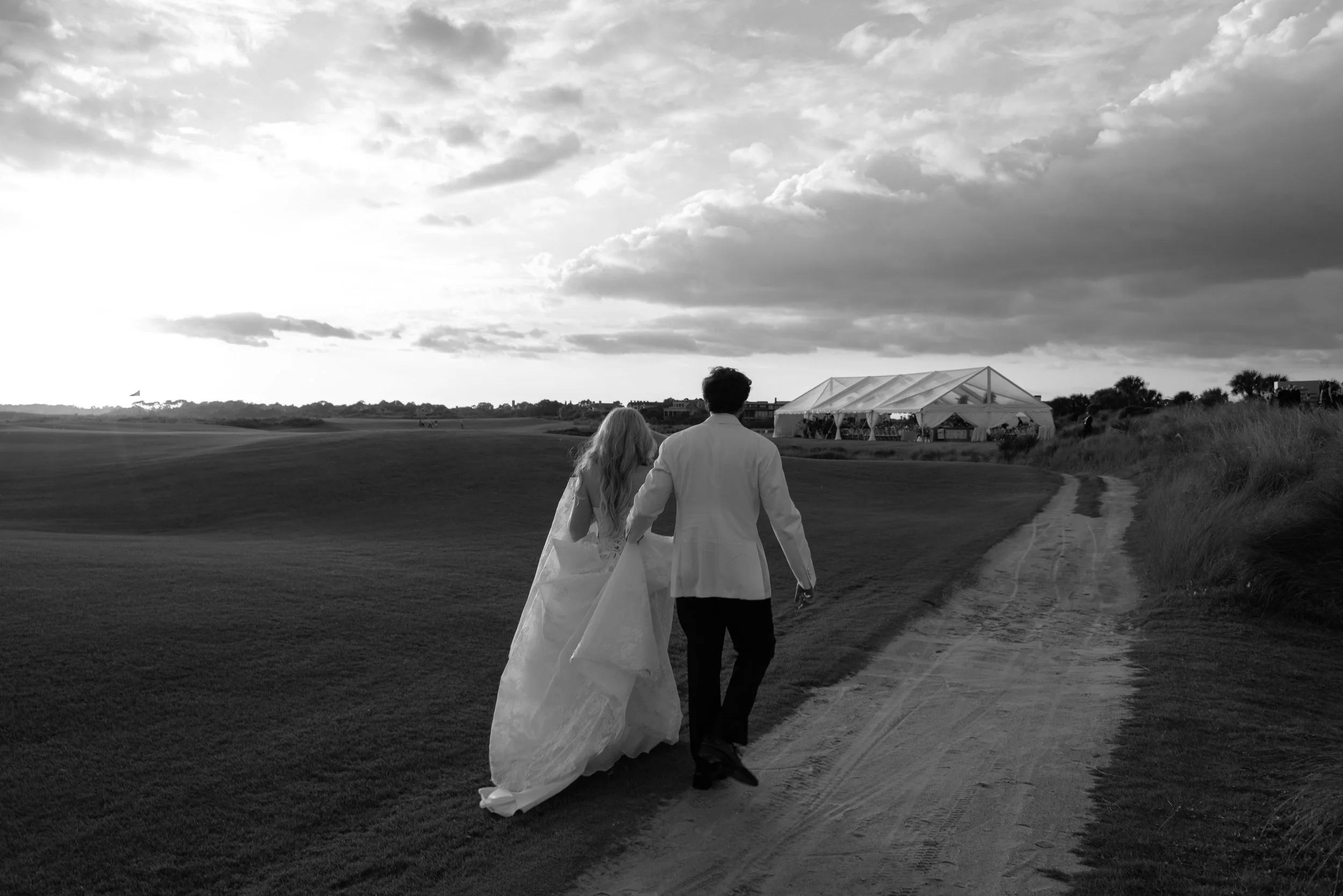 Katie and Trevor walk toward their reception tent along a quiet Ocean Course path in a black-and-white landscape photograph.
