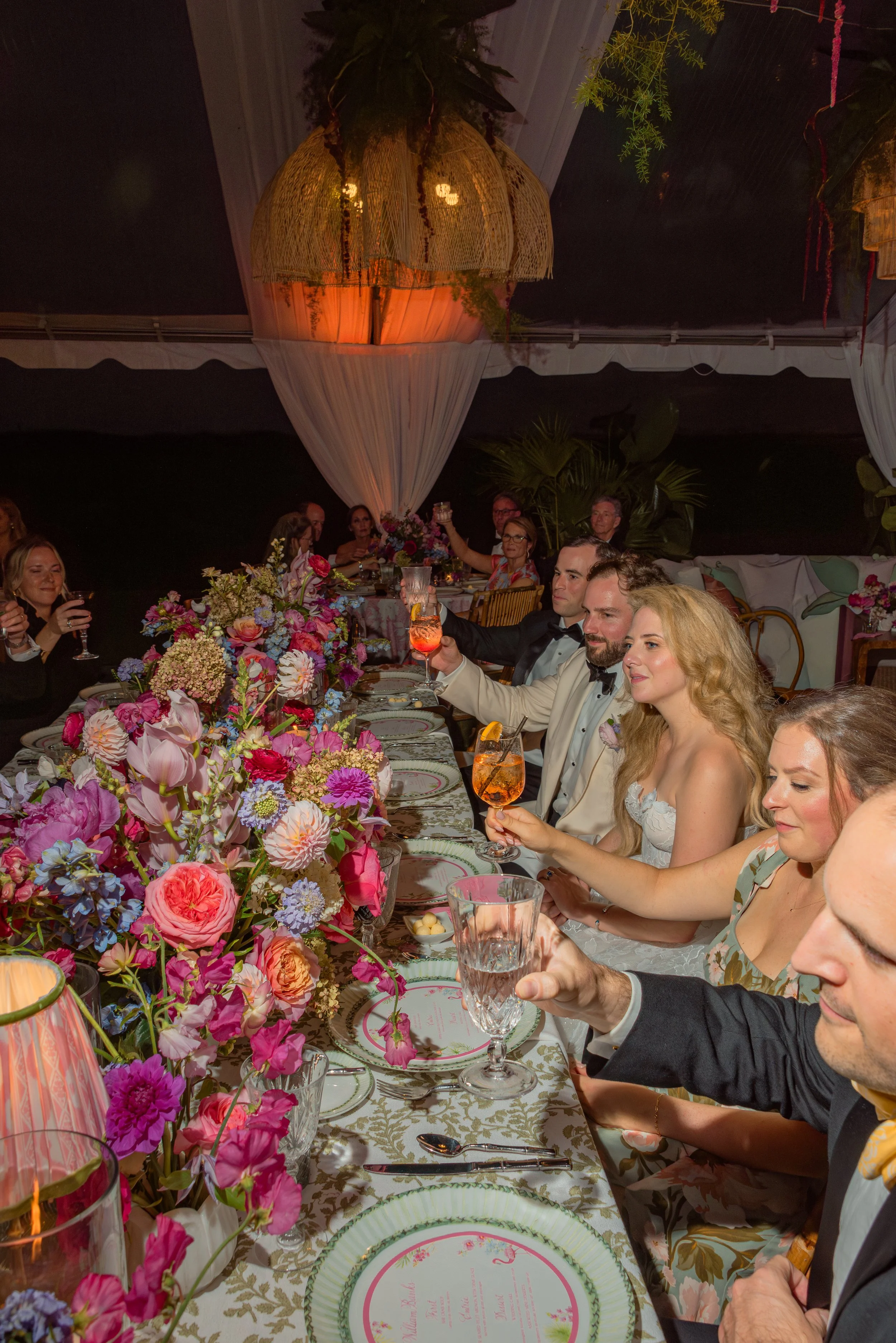 Katie, Trevor, and their guests raise glasses around a long, densely flowered reception table.