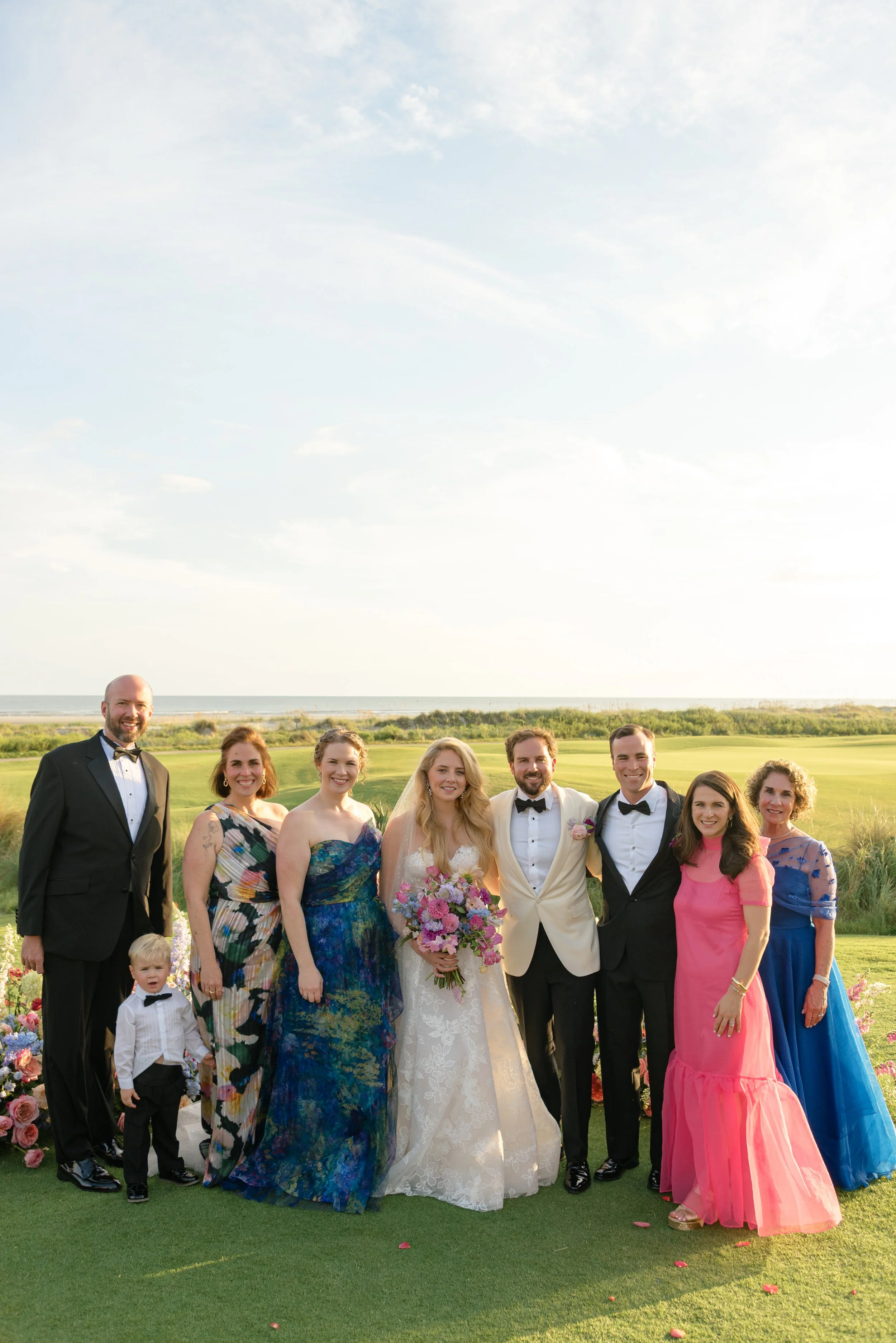 Katie and Trevor pose with their families in colorful formal attire against the coastal landscape at The Ocean Course on Kiawah Island.
