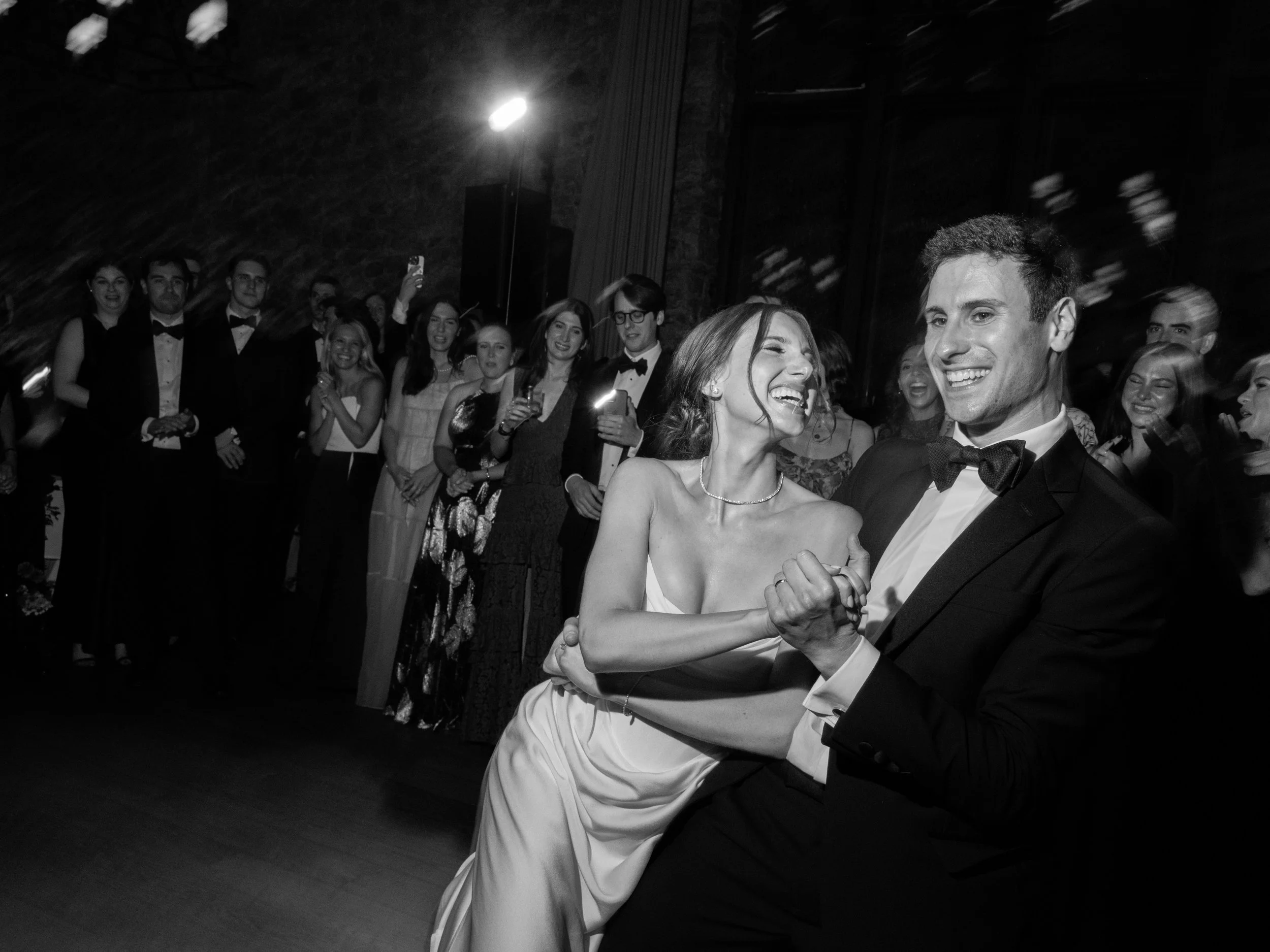 Black-and-white candid dancing photo of Will and Jennie smiling among family and friends during the reception.
