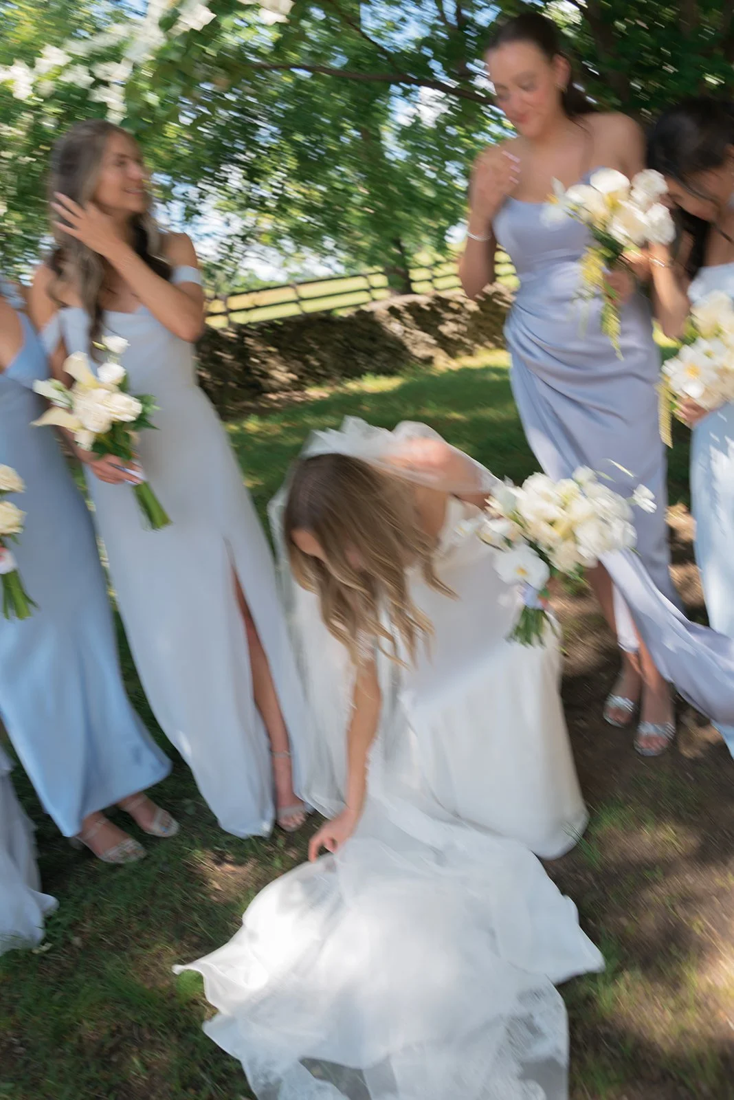 Bride bends down to fix the train of her wedding gown during formal photos with her bridesmaids