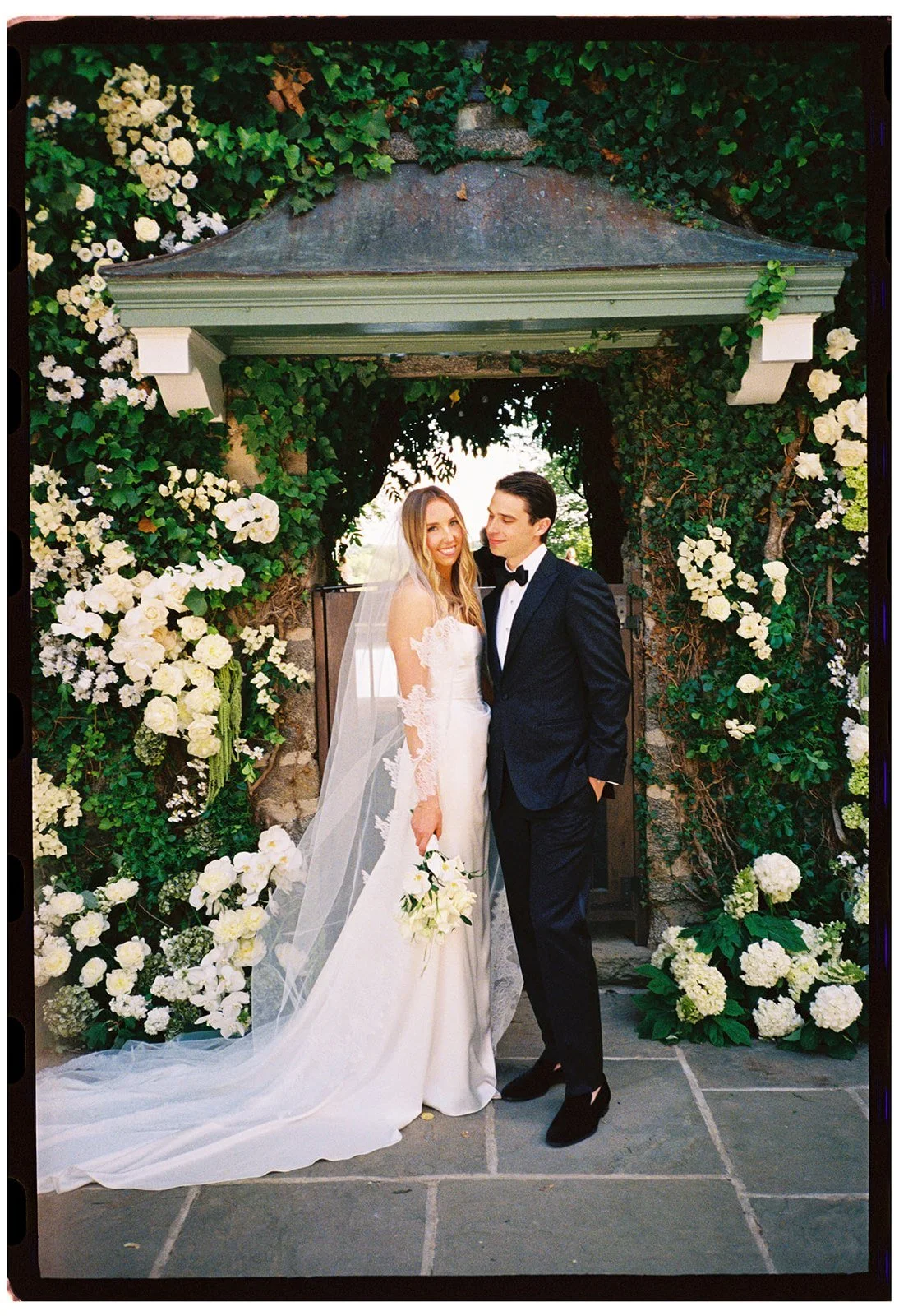 Full-length film portrait of the bride and groom standing beneath the floral Ivy Wall at Goodstone Inn.