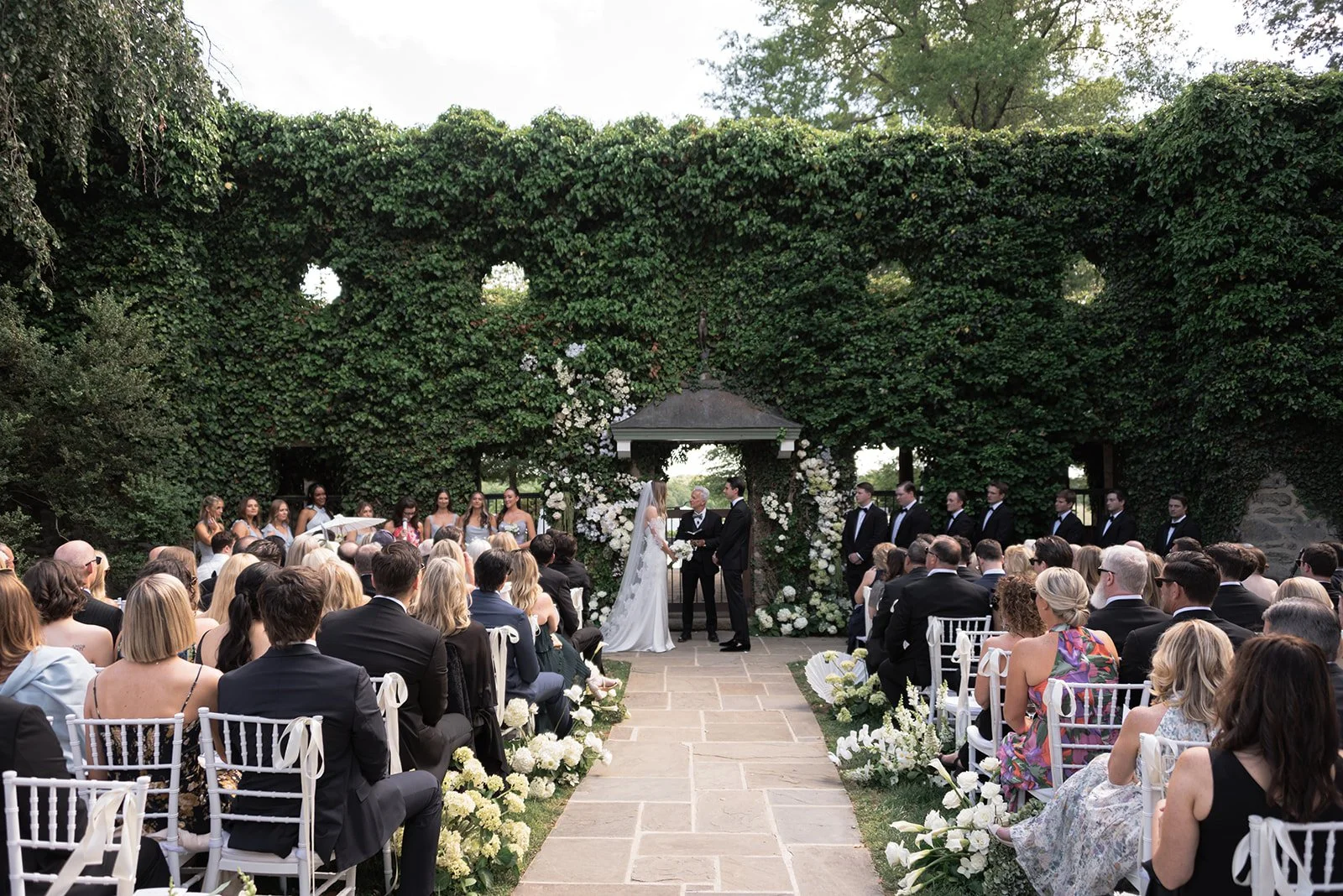 Wide view of an outdoor wedding ceremony at the Ivy Wall at Goodstone Inn with guests seated in garden rows.