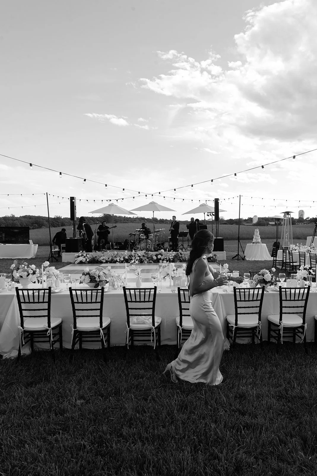 Black-and-white wide view of long dinner tables arranged for an outdoor reception in the Hayfield at Goodstone Inn with a bridesmaid in a silk dress walking next to the tabe. 