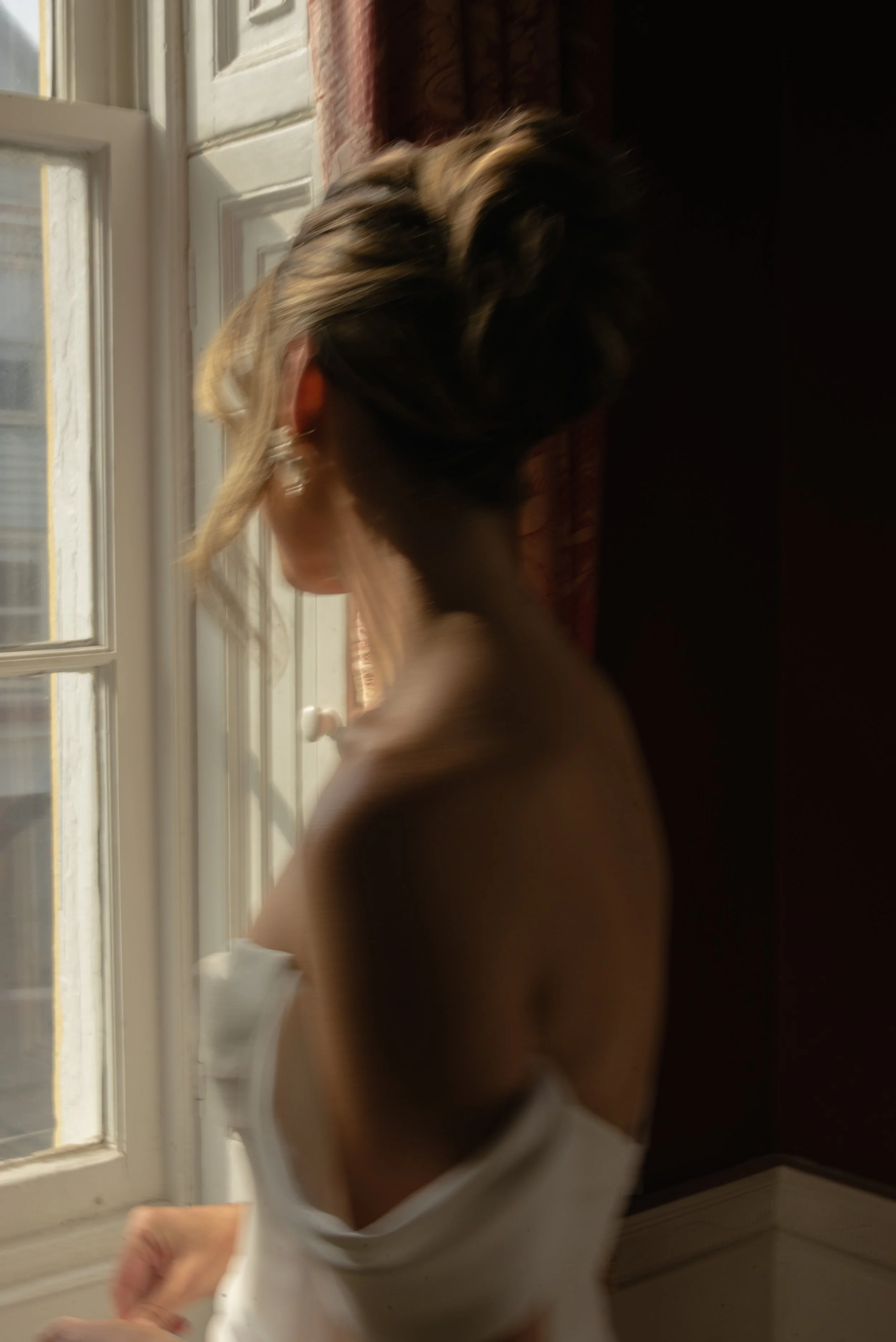 Soft-focus bridal portrait of a bride standing near a window, capturing a quiet moment of reflection before her wedding ceremony.