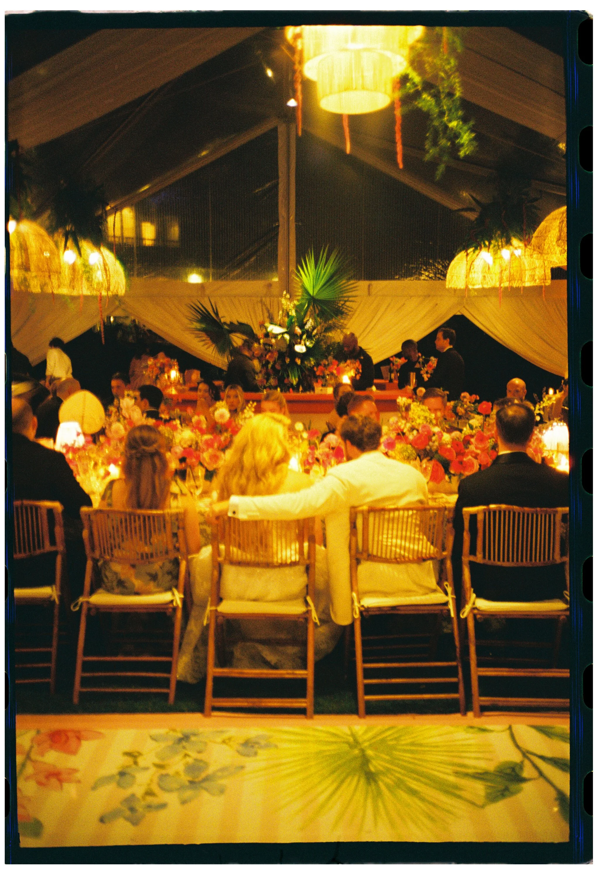 Film photo of guests dining beneath warm chandeliers and tropical greenery inside the tent.