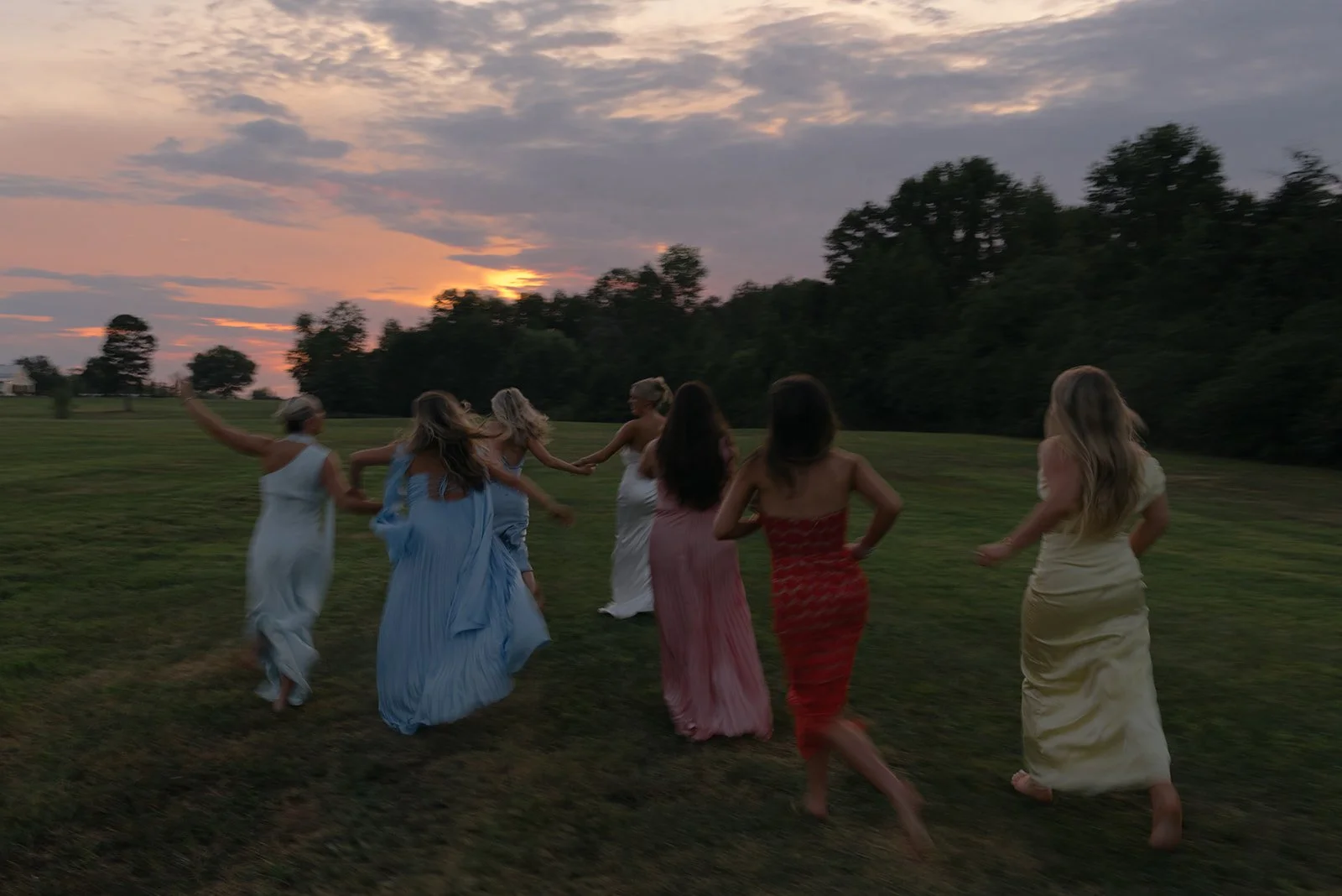 Bride and bridesmaids running together across a grassy field during a destination wedding celebration at sunset.