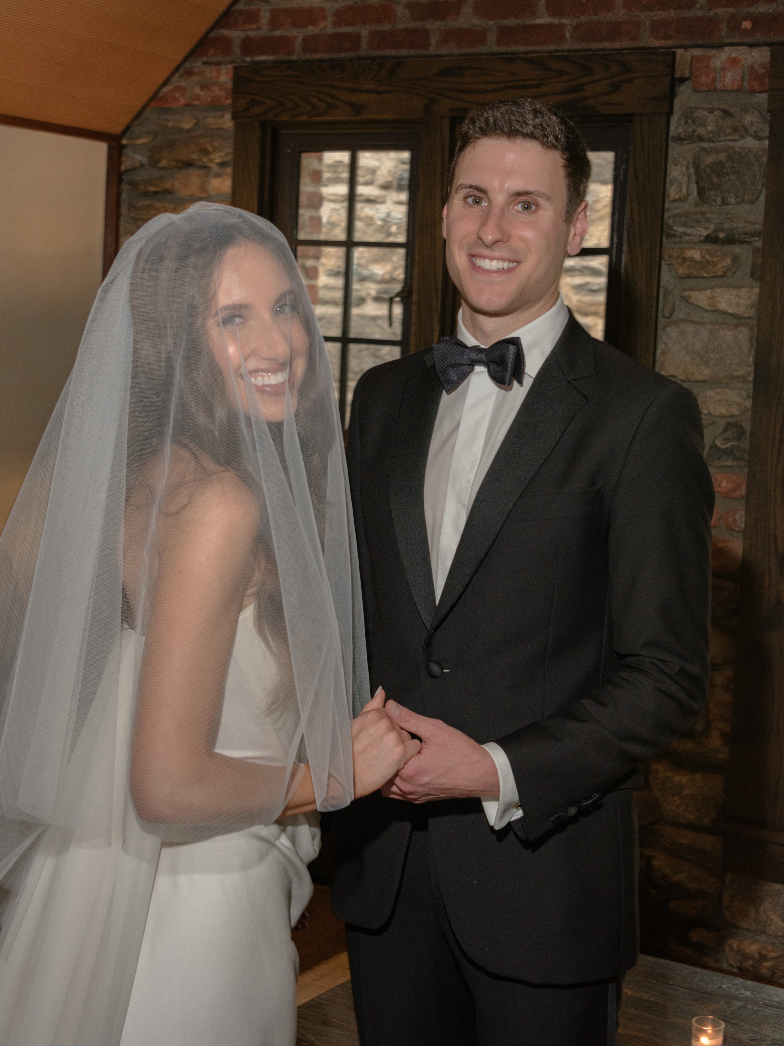 The bride wears a blusher veil during the Ketubah ceremony and smiles brightly with her groom while they hold hands.