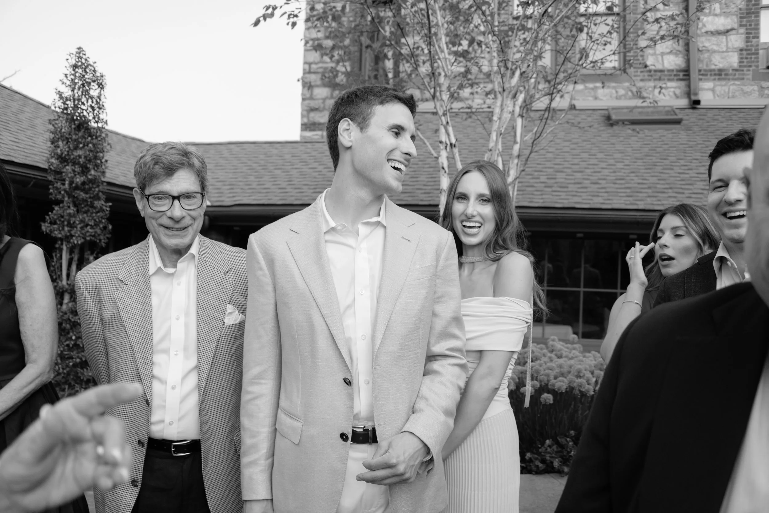 Black-and-white candid of groom laughing with family and friends during the rehearsal dinner.