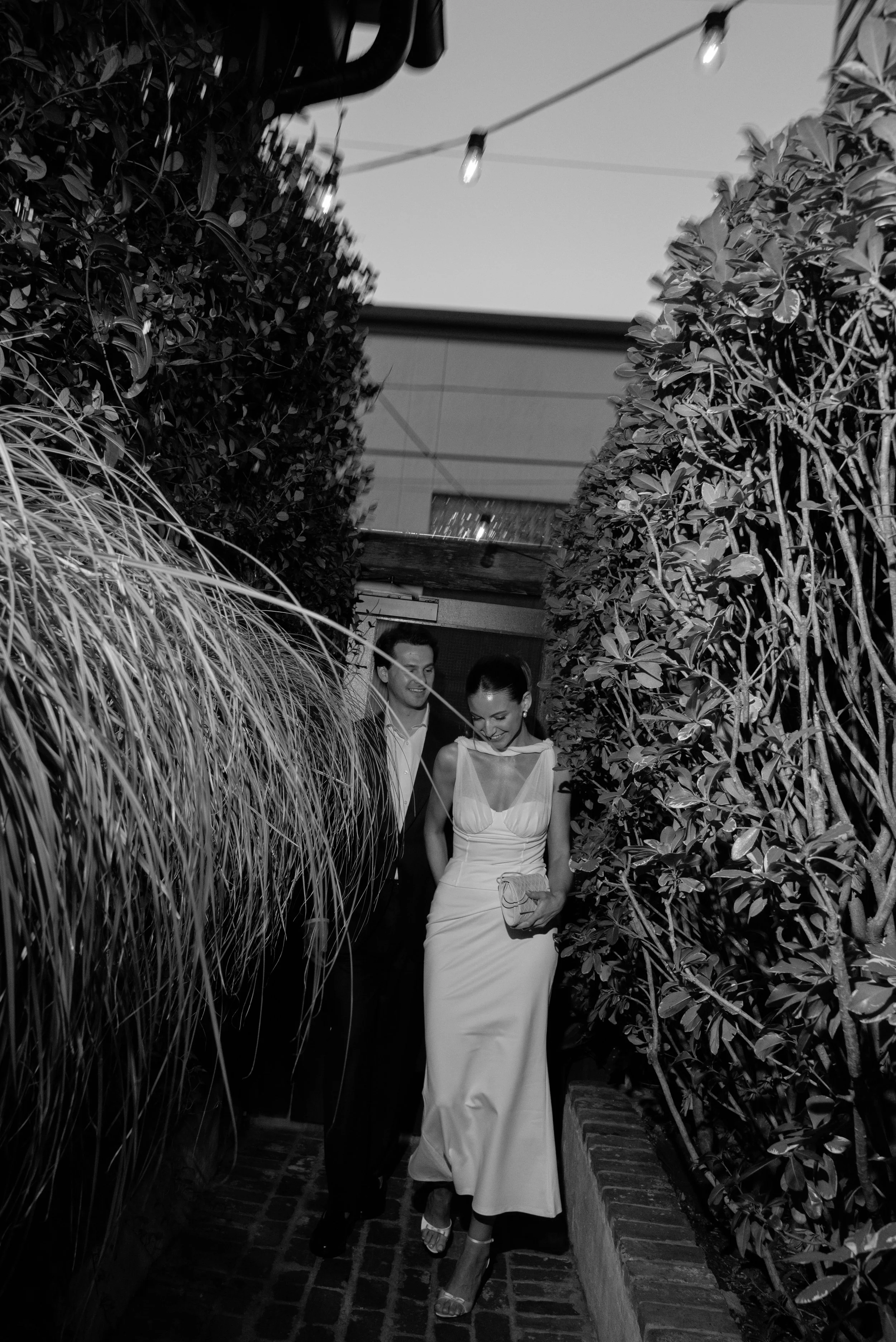 Black and white image of the bride and groom entering their rehearsal dinner location at Leon's Oyster Shop in Charleston, South Carolina, among lush greenery and market lights overhead. 