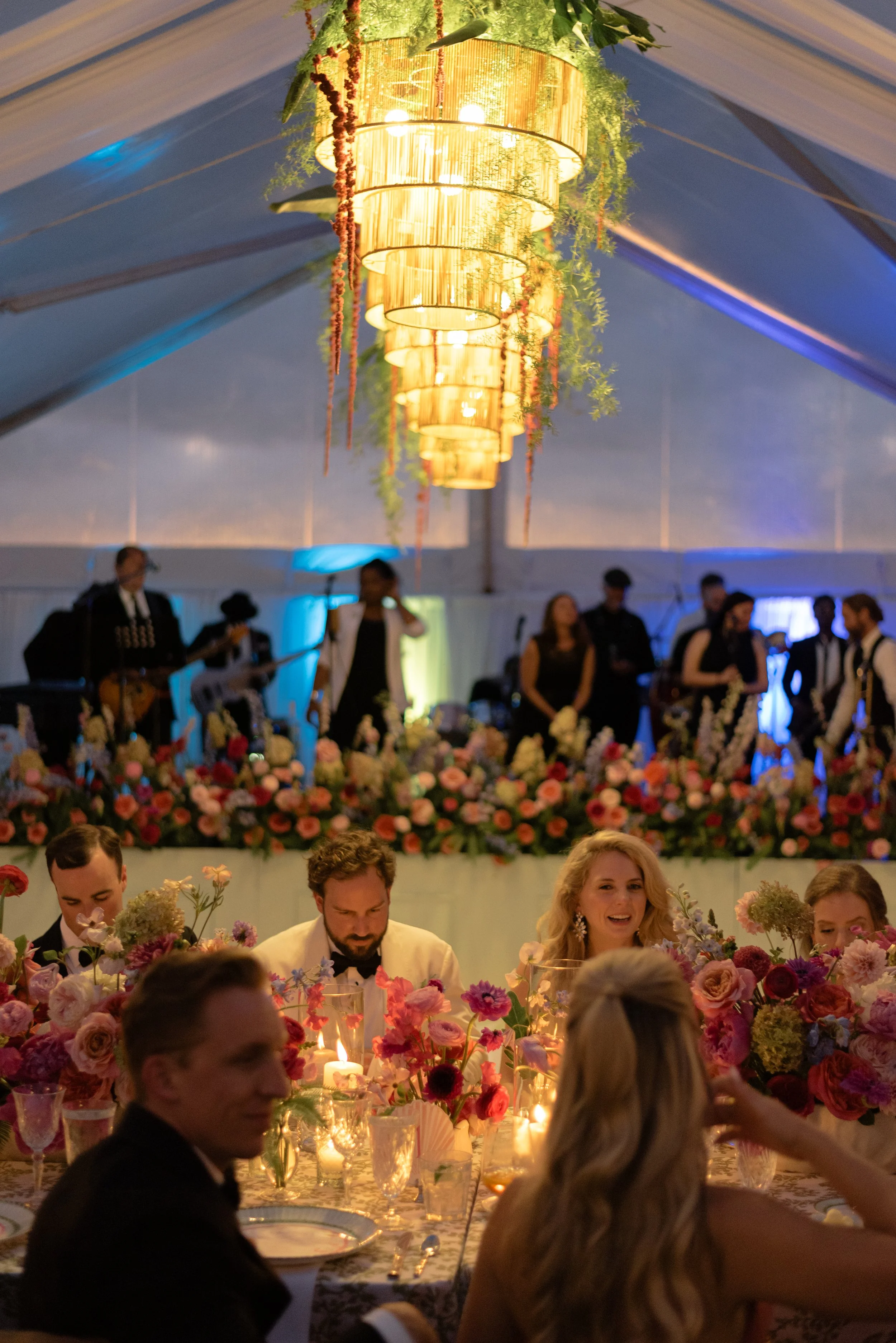 Guests dine beneath glowing woven chandeliers surrounded by colorful flowers inside the Ocean Course reception tent.