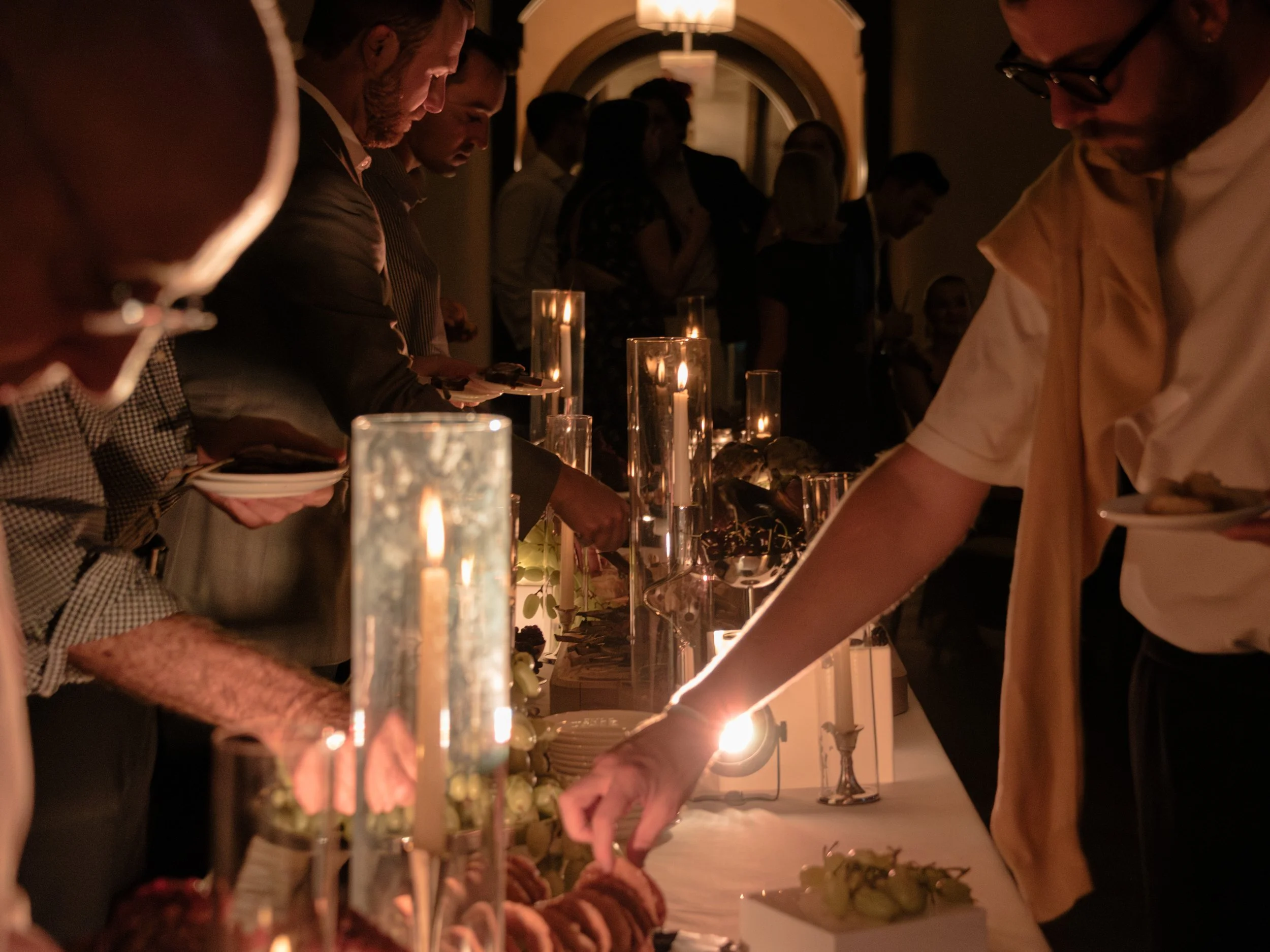 Guests reaching across the candlelit food display during the lively rehearsal dinner.