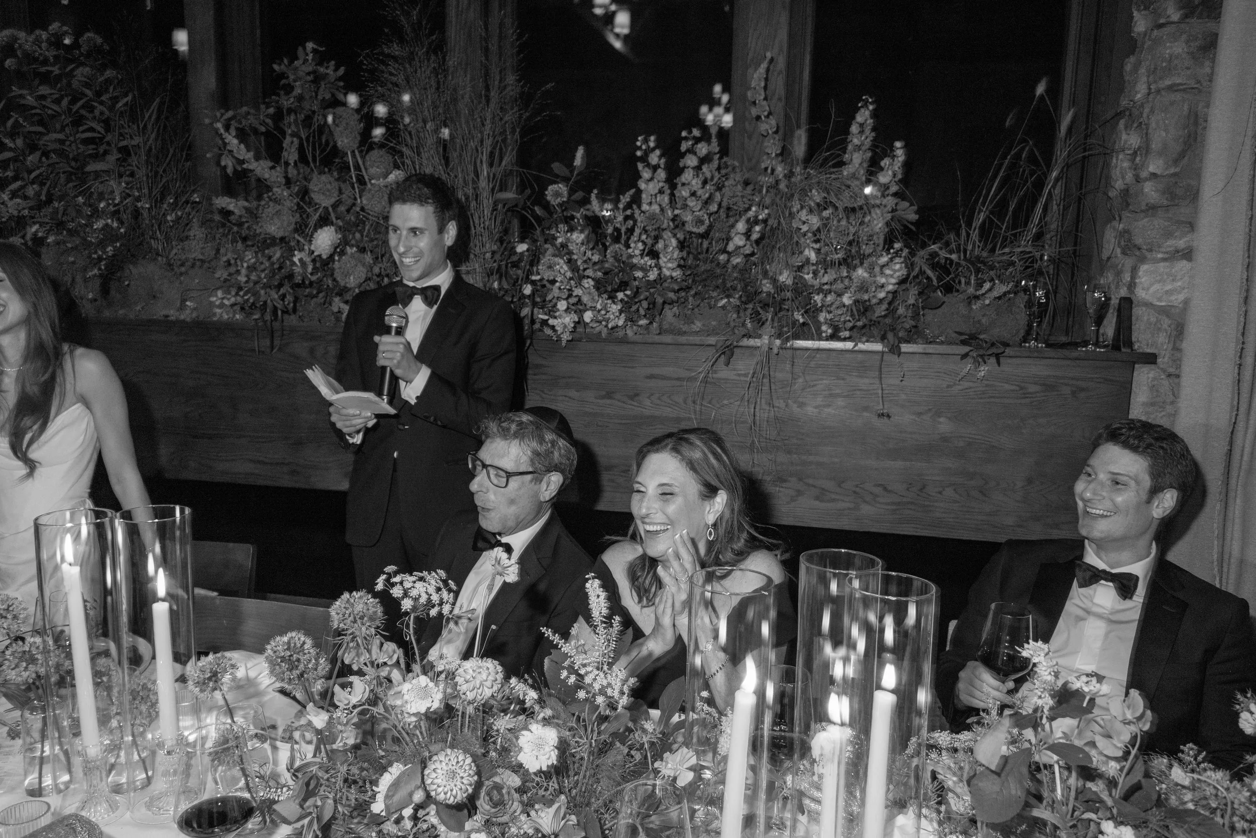 Black-and-white view of the candlelit reception table framed by floral design and historic large windows and stone walls.