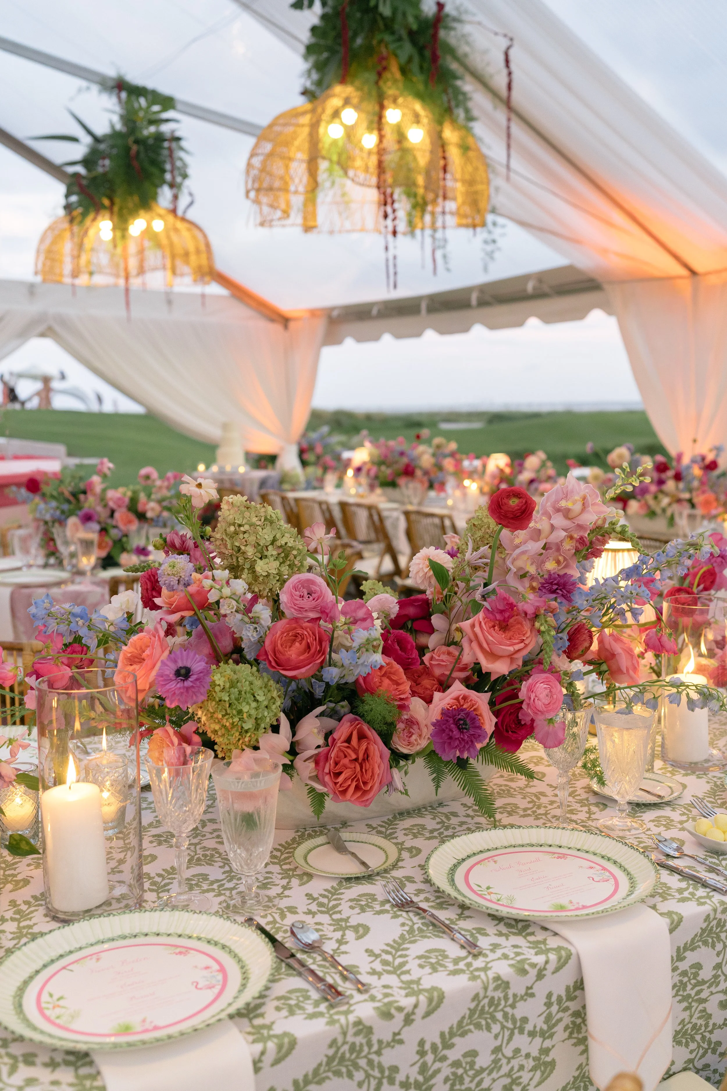 Colorful wedding tablescape features layered flowers, patterned linens, pink plates, candles, and woven chandeliers.