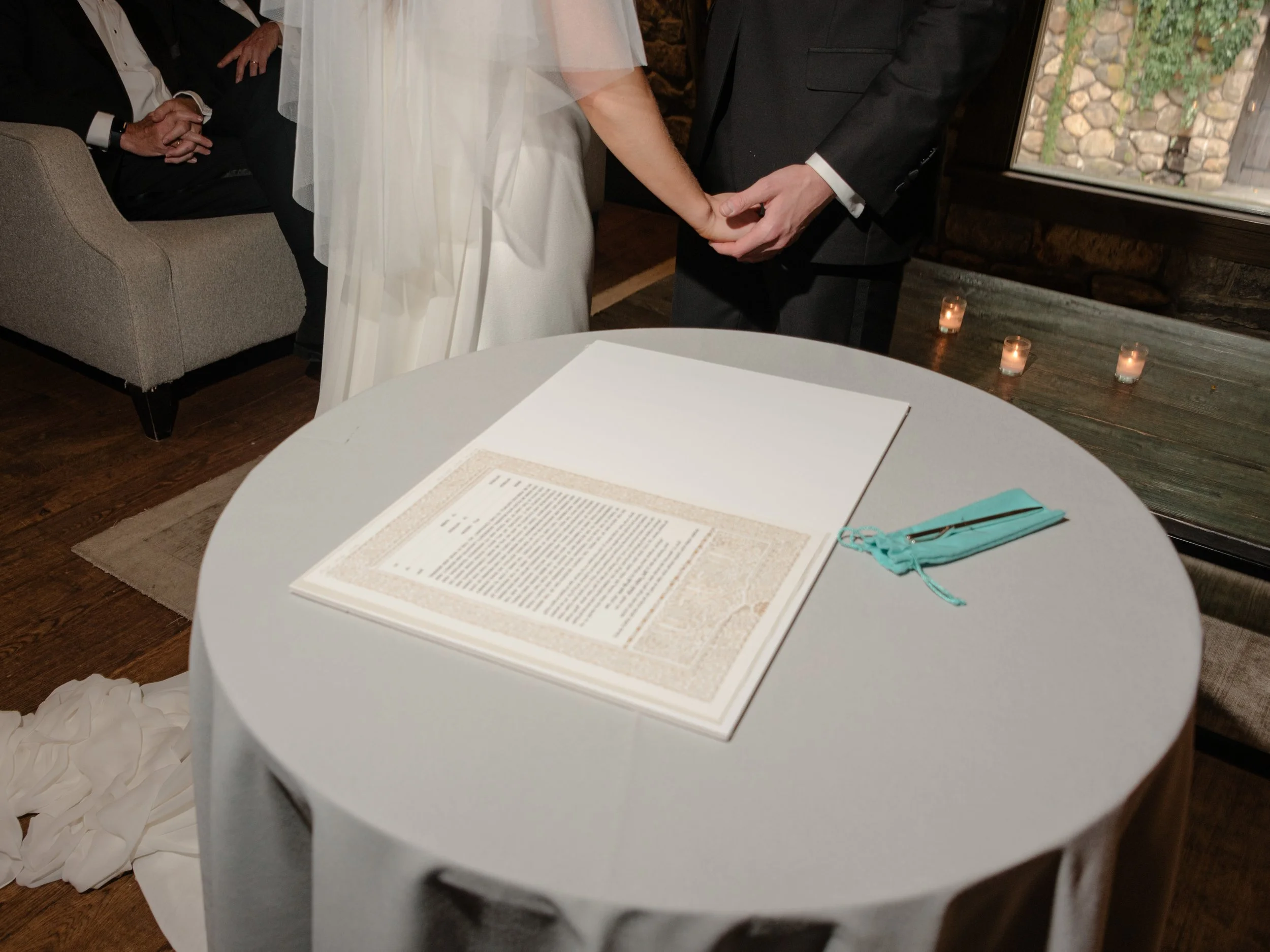 The couple holds hands before sitting at the table where their Ketubah is waiting to be signed and can be seen here on the table beneath their clasped hands. 