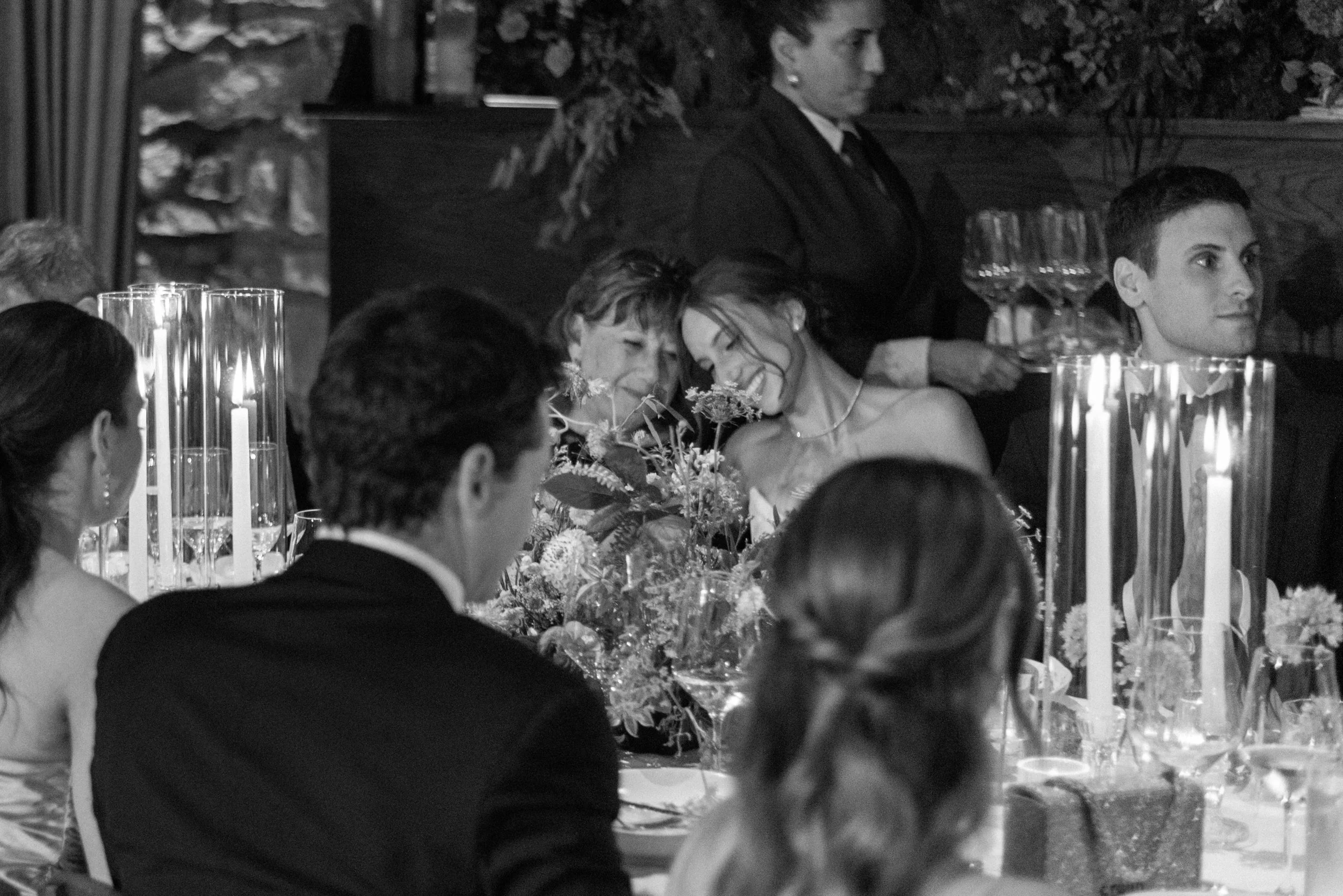 Black-and-white dinner scene of the bride and her mother sharing a sweet, quiet moment among candles and floral arrangements.