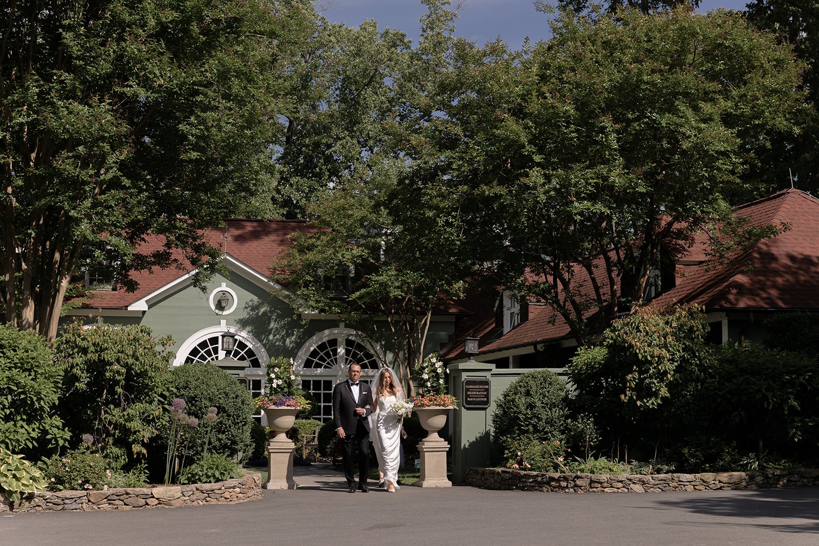 Wide view of Goodstone Inn gardens and ceremony area framed by mature trees and estate architecture as the bride and her father make their entrance to the ceremony.