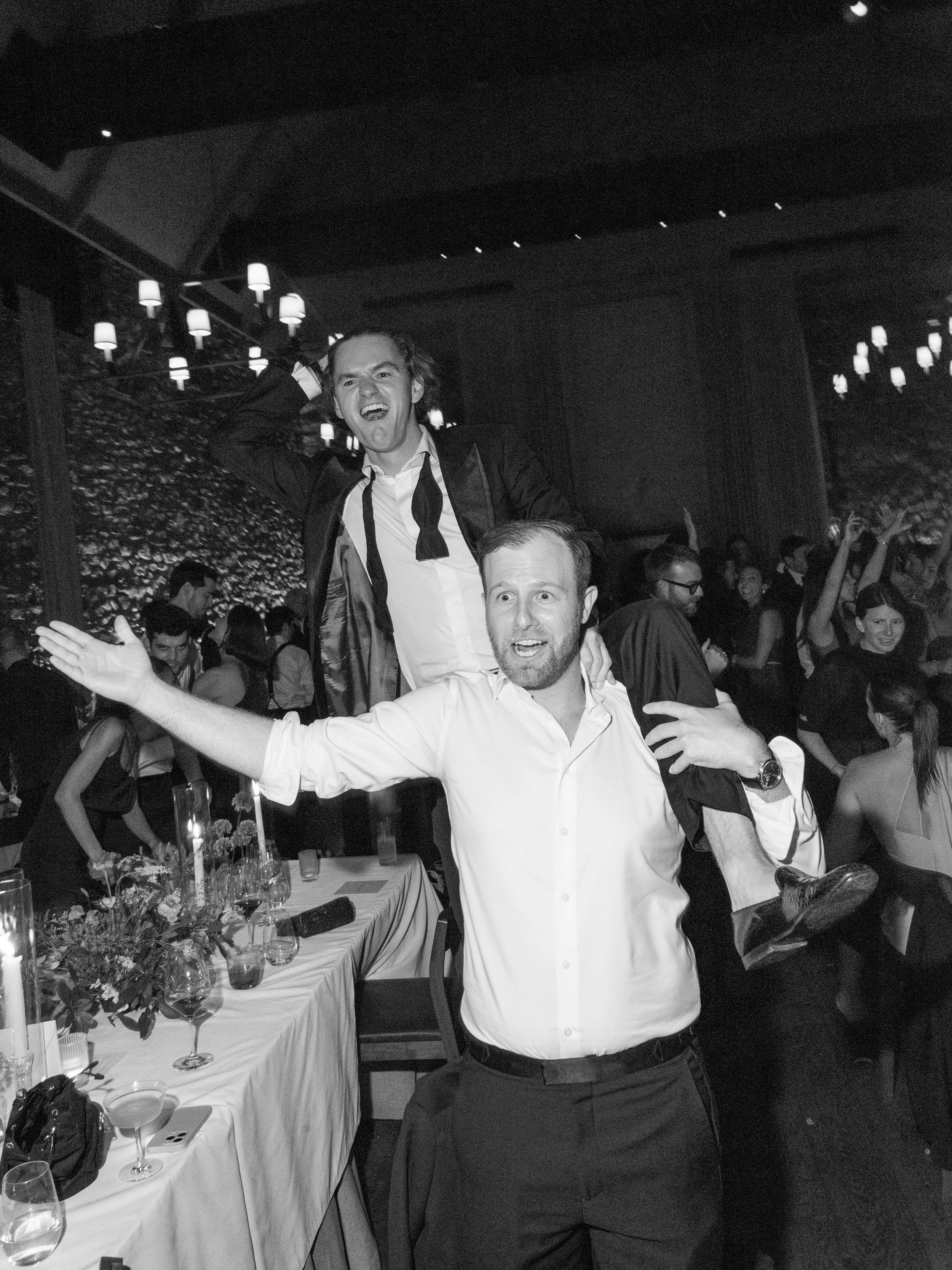 Black-and-white candid of a guest dancing enthusiastically on the shoulders of another guest during the late-night reception.