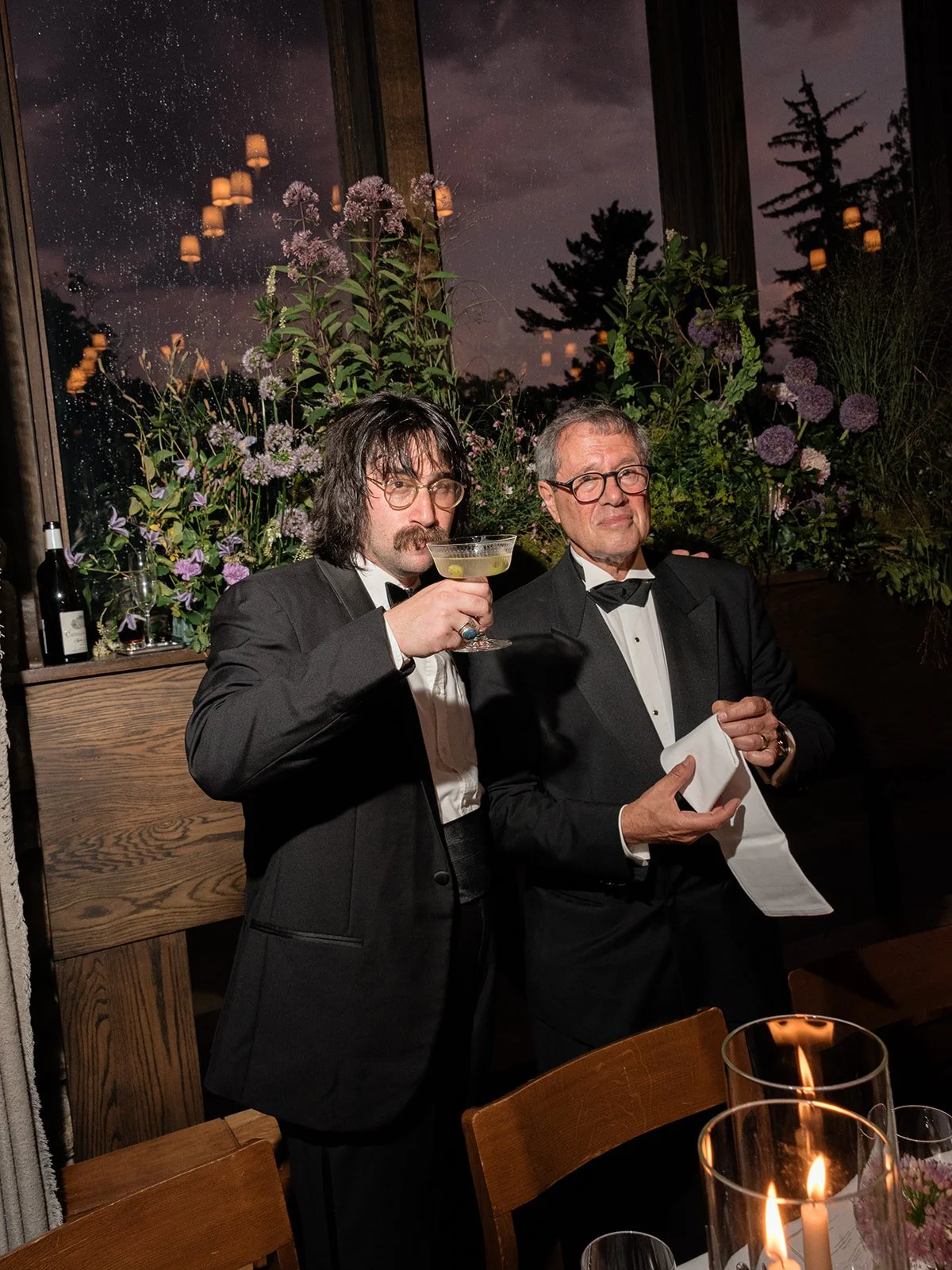 Two wedding guests in black-tie attire share a drink beside a candlelit floral display during the reception.