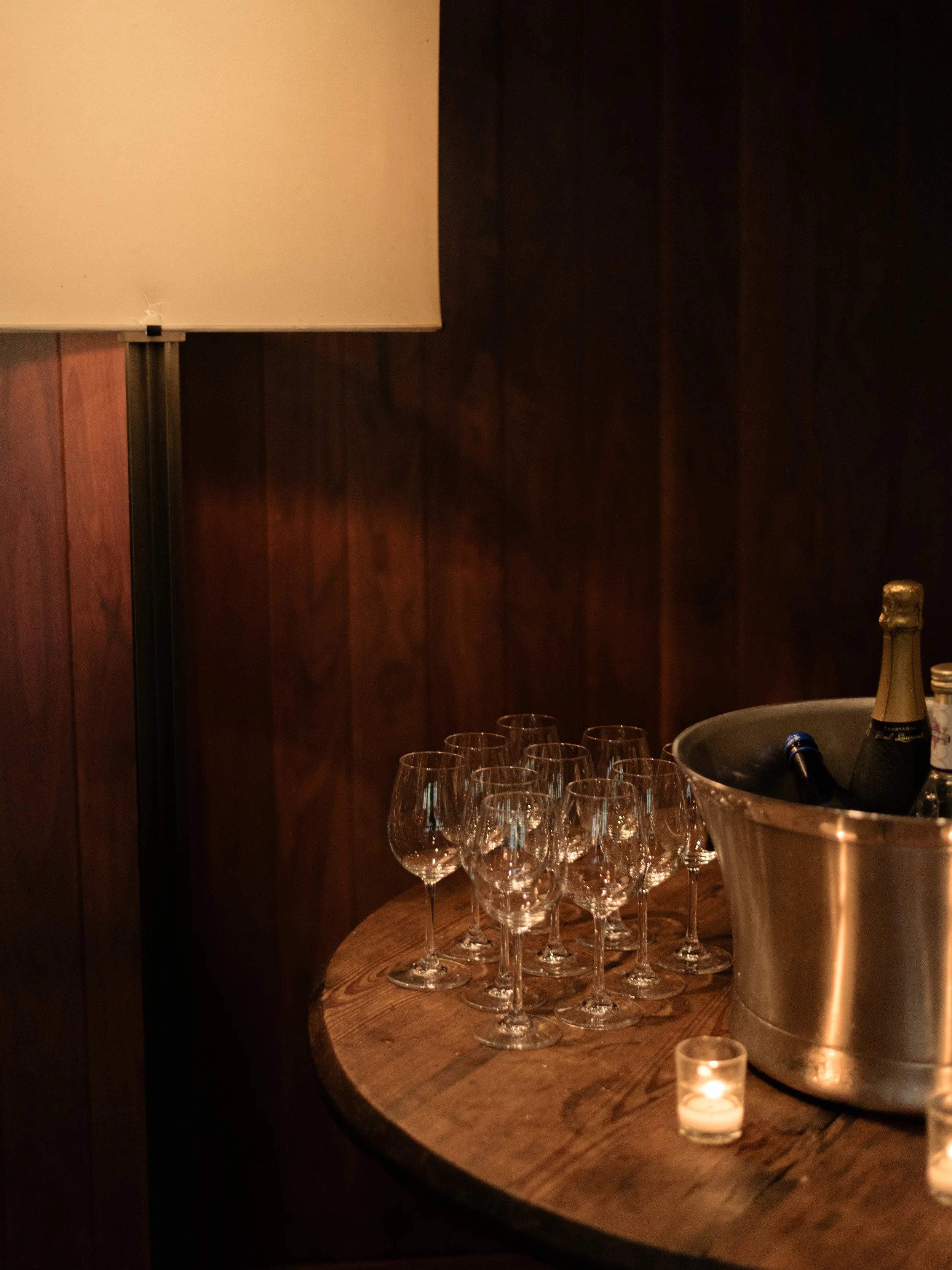 An ambient design photograph of wine glasses and silver champagne buckets positioned on a round wooden table with a table lamp set against rich warm wood walls at Blue Hill at Stone Barns.
