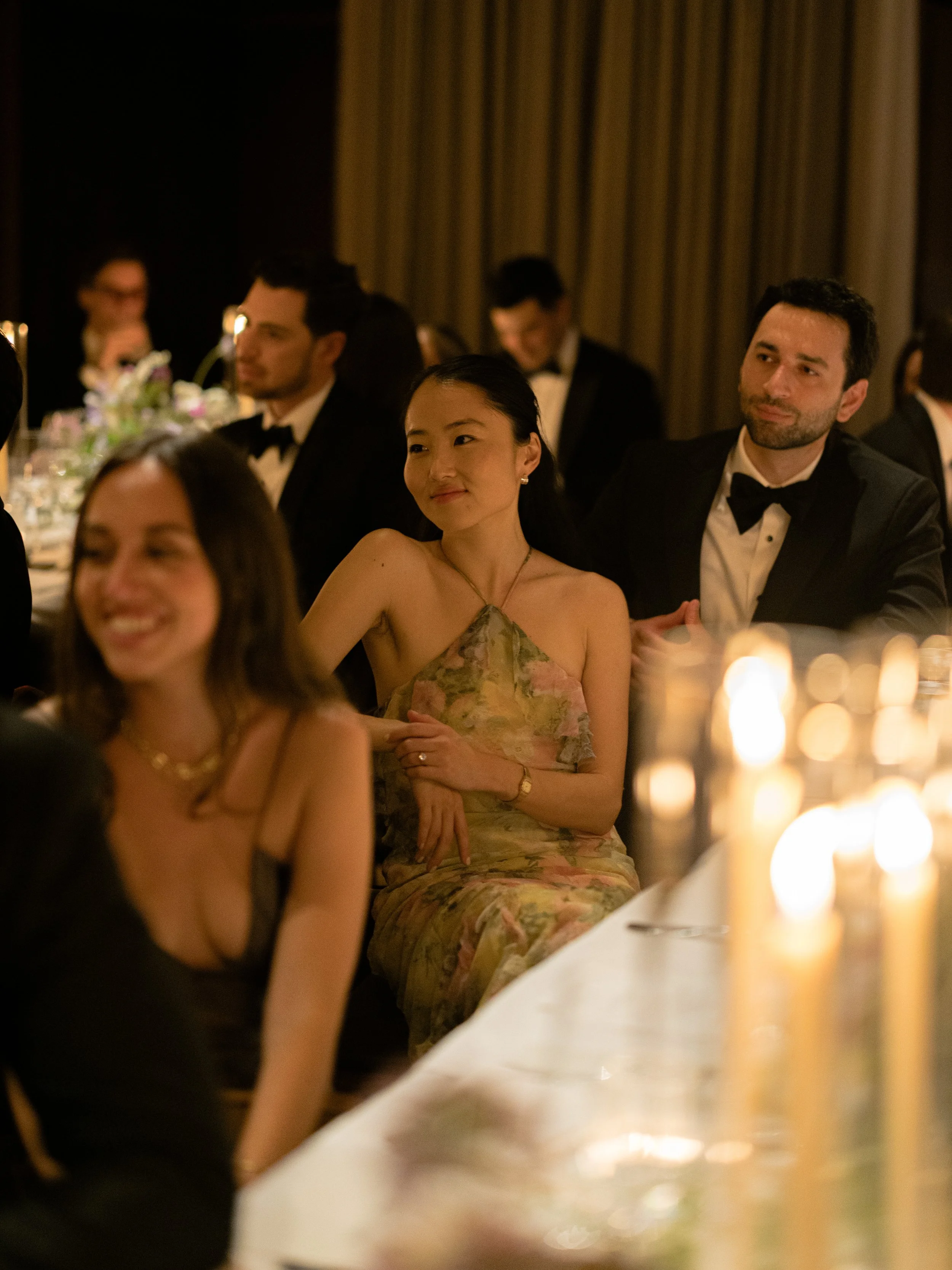 Guests listening during speeches surrounded by taper candles and floral arrangements at the reception.