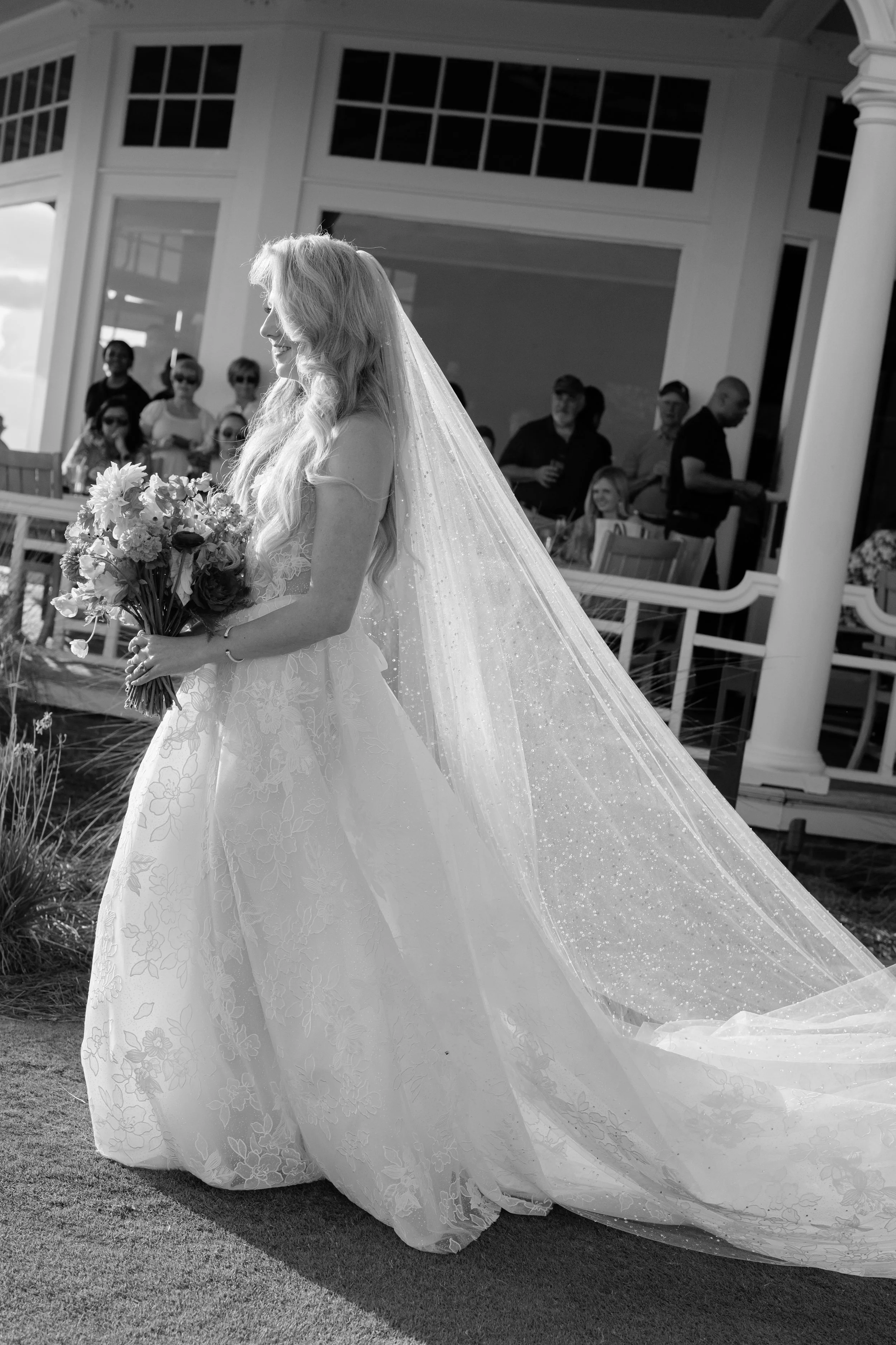 Black-and-white photograph of Katie entering the outdoor ceremony in her lace Galia Lahav gown and long veil.