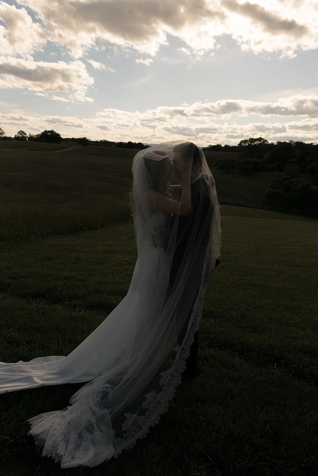 Bride and groom embrace beneath the cathedral veil in a golden field beneath a dramatic sky