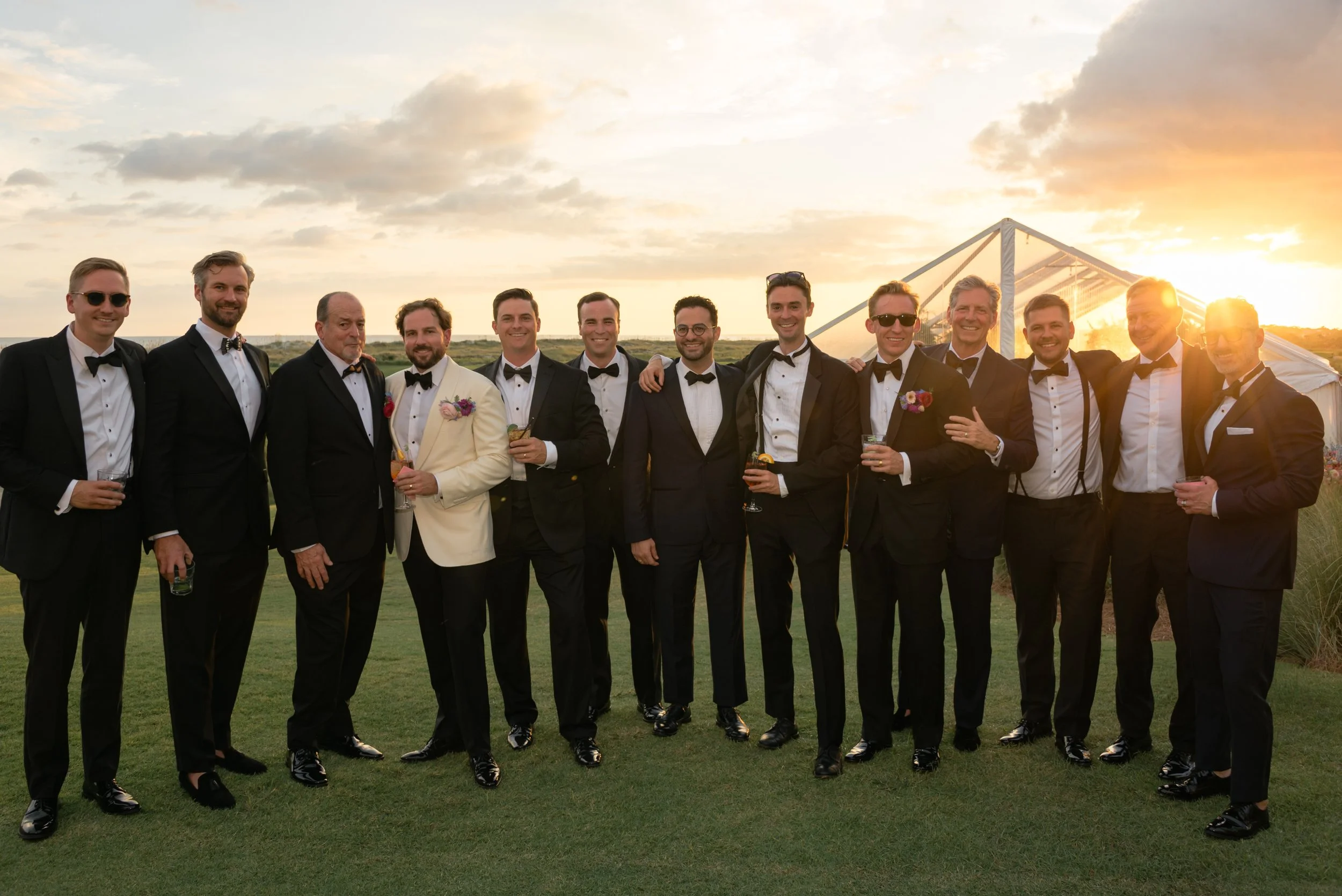 Trevor and his groomsmen pose in black-tie attire with the glowing reception tent and sunset behind them.