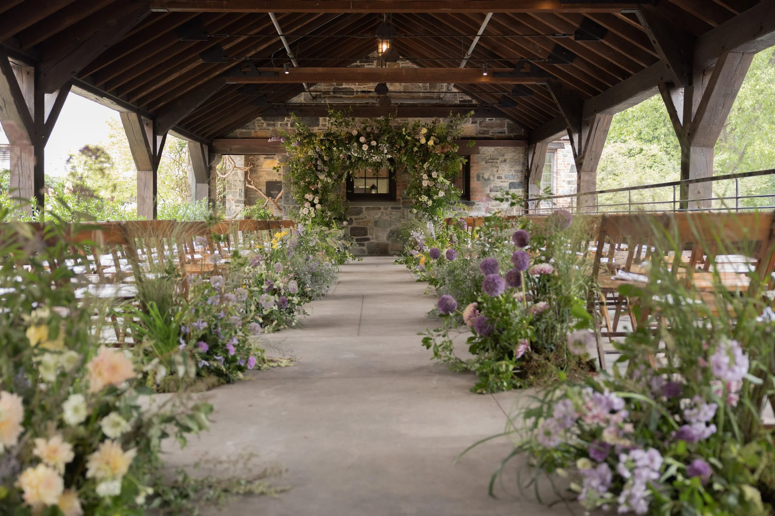 The pergola in the courtyard of Blue Hill at Stone Barns filled with whimsical floral designs in shades of purple and lilac and butter yellow designed by Wild and Birch. 