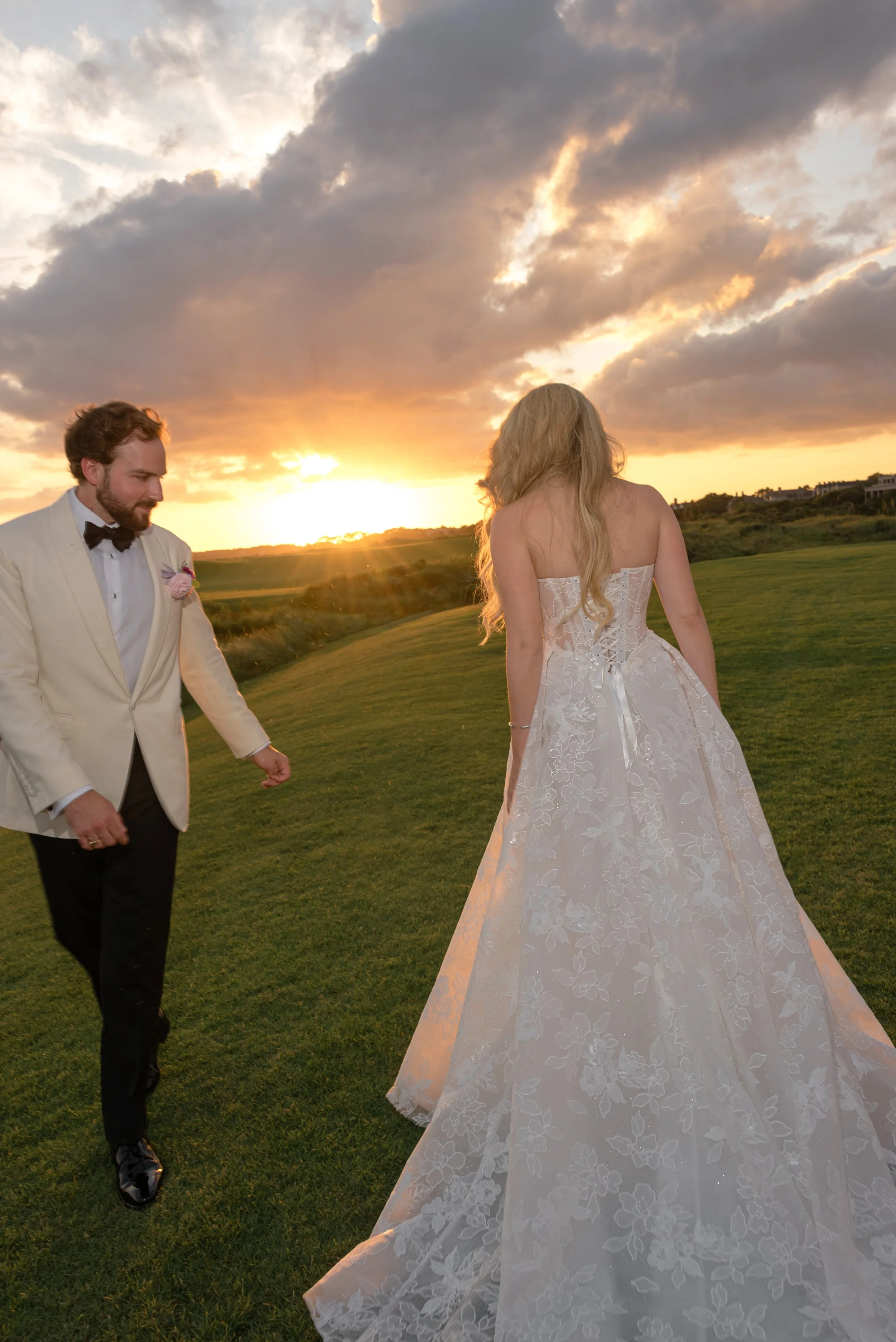 The bride and groom walk together across the golf course as the sun sets behind them.