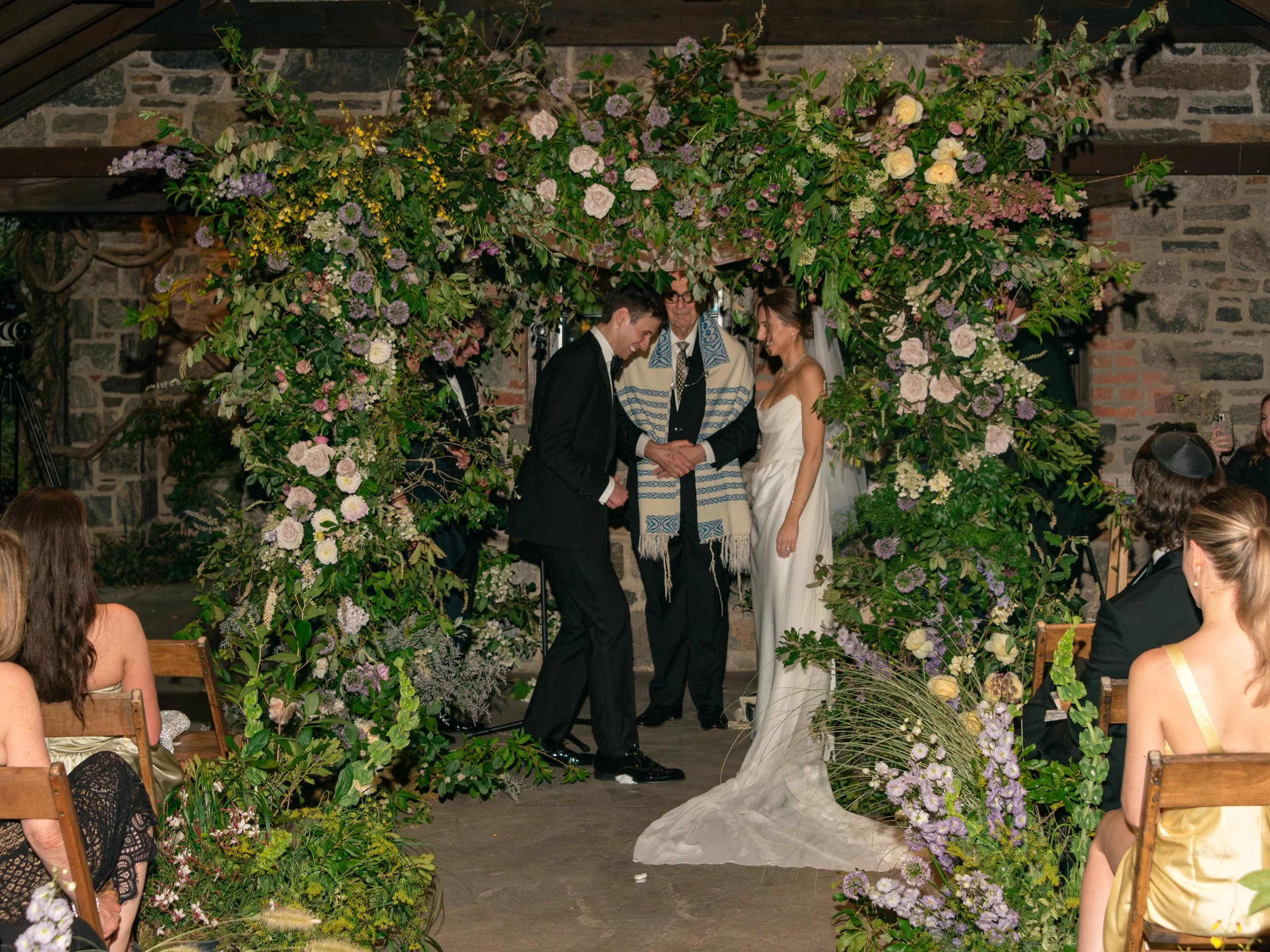 Guests gather for a Jewish wedding ceremony surrounded by floral design and historic stone architecture.
