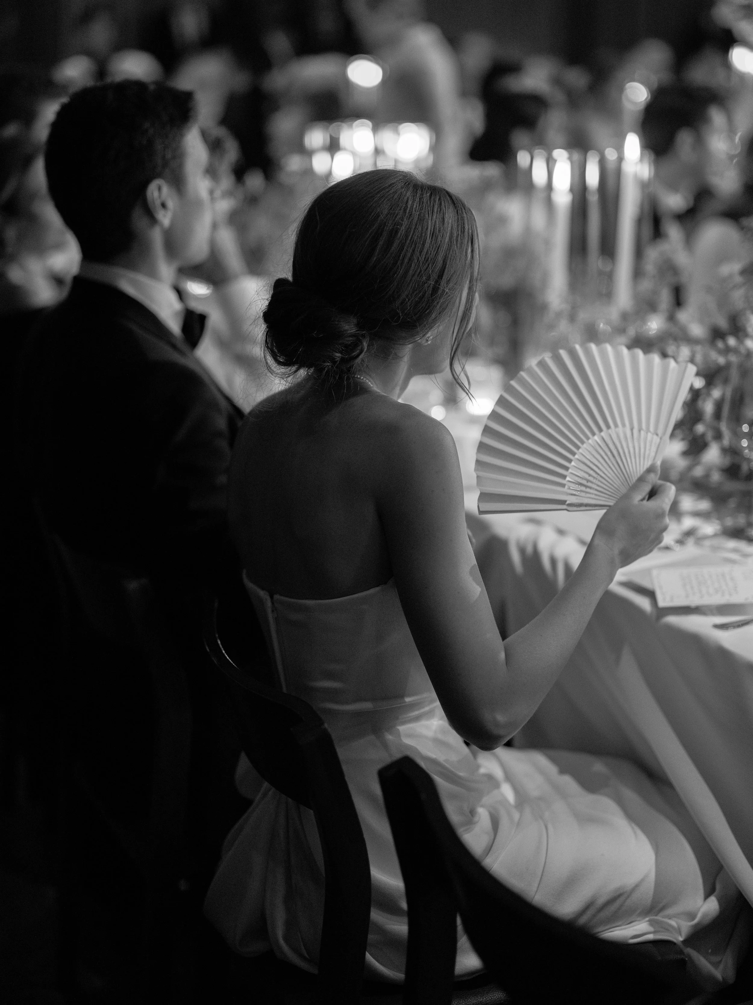 Black-and-white candid of the bride holding a hand fan during the candlelit wedding dinner.