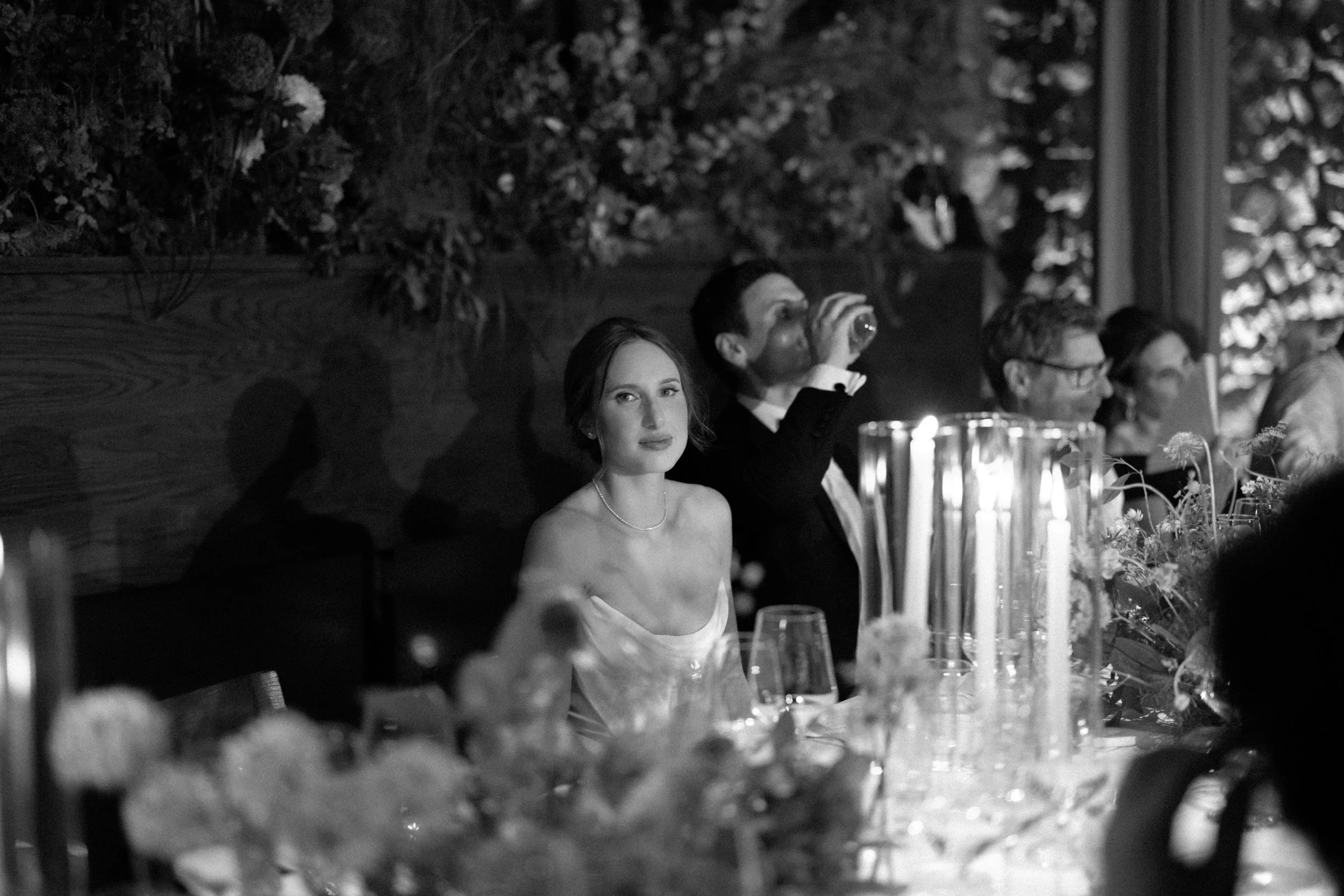 Black-and-white candid portrait of the bride framed by candlelight and flowers during dinner at Blue Hill at Stone Barns.