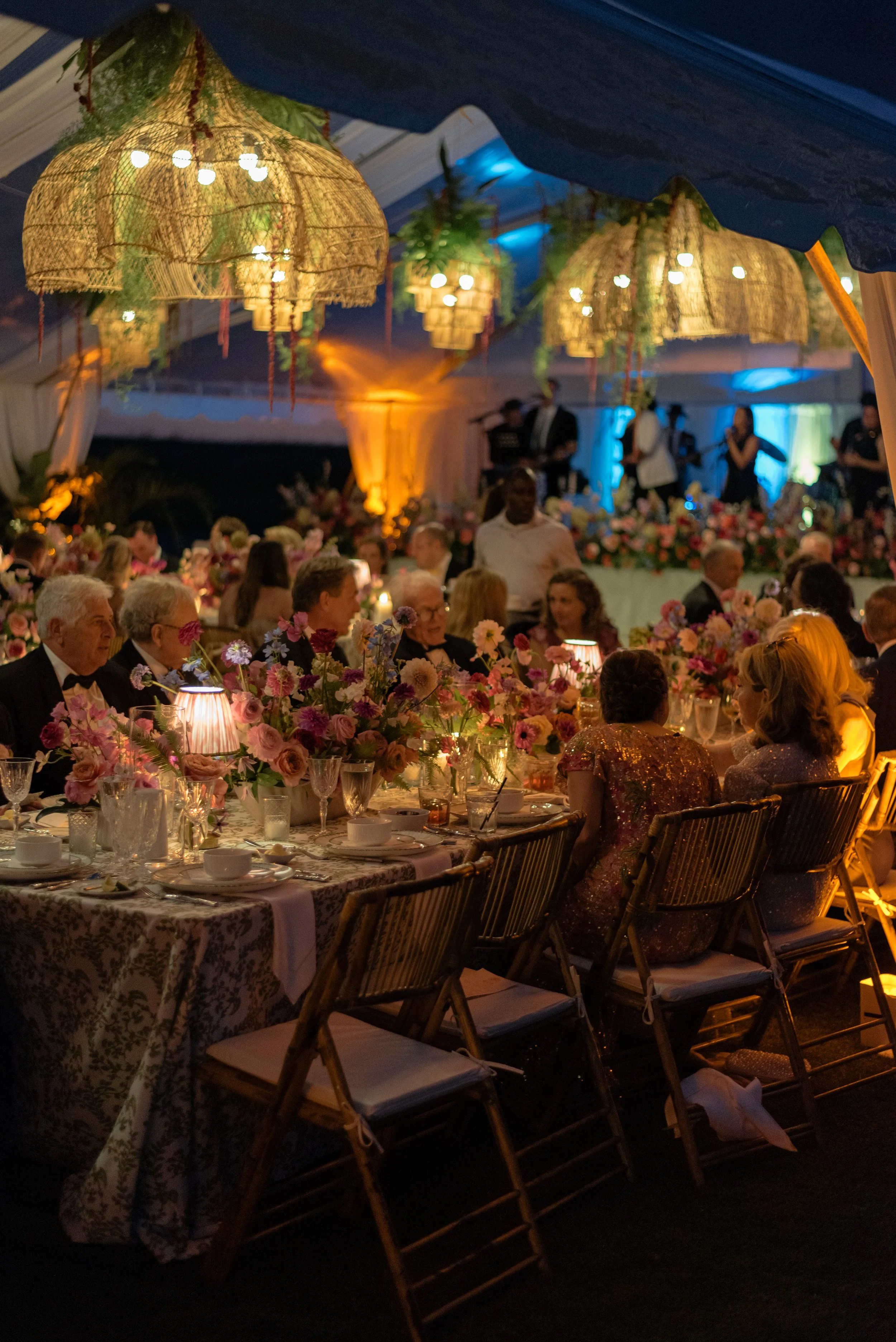 Guests dine beneath glowing woven chandeliers and colorful flowers inside the clear-top wedding tent.