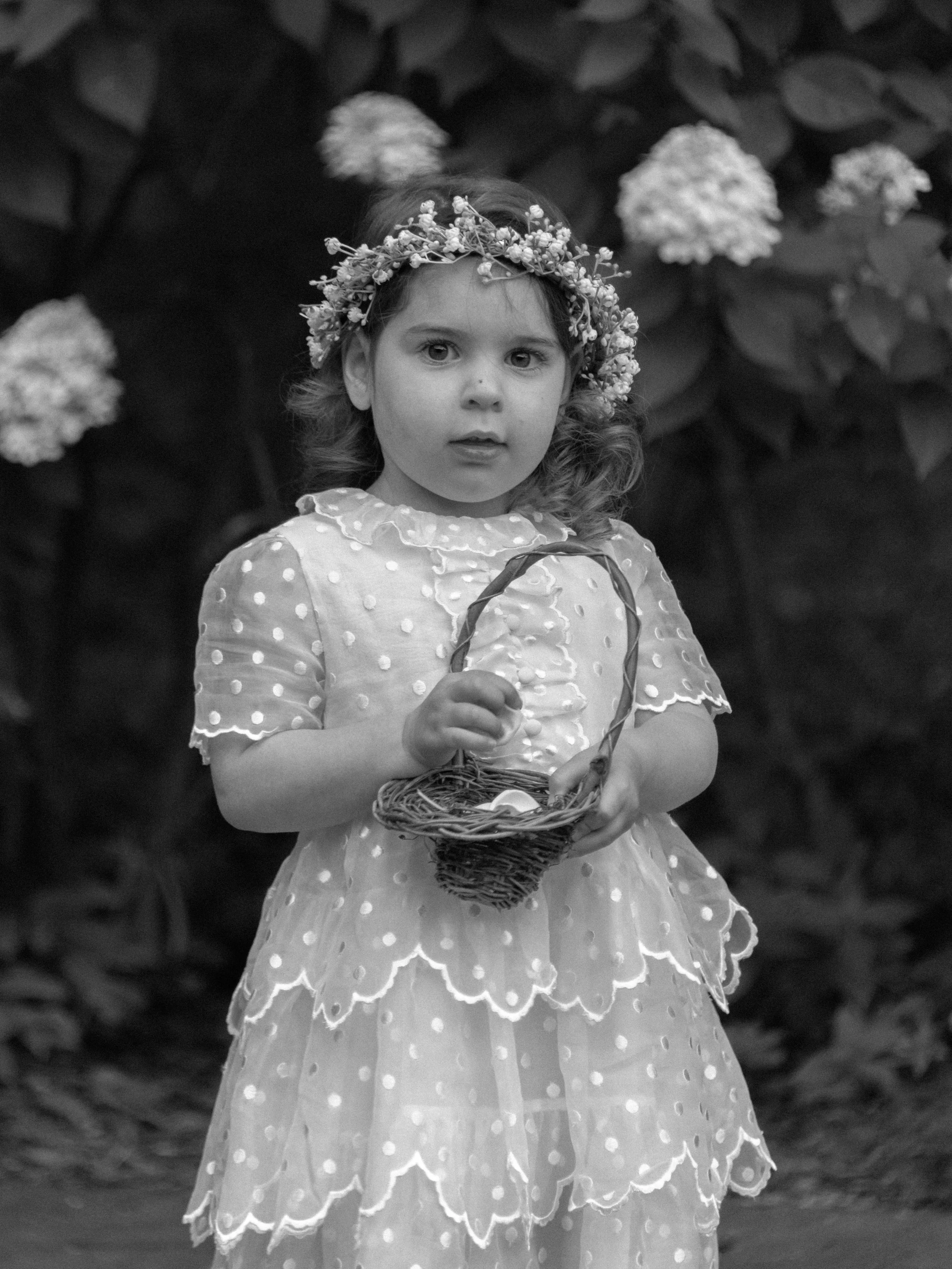 The flower girl dressed in layer of laces of wearing a baby's breath floral crown carries a small wicker basket of flower petals as she enters the wedding ceremony.