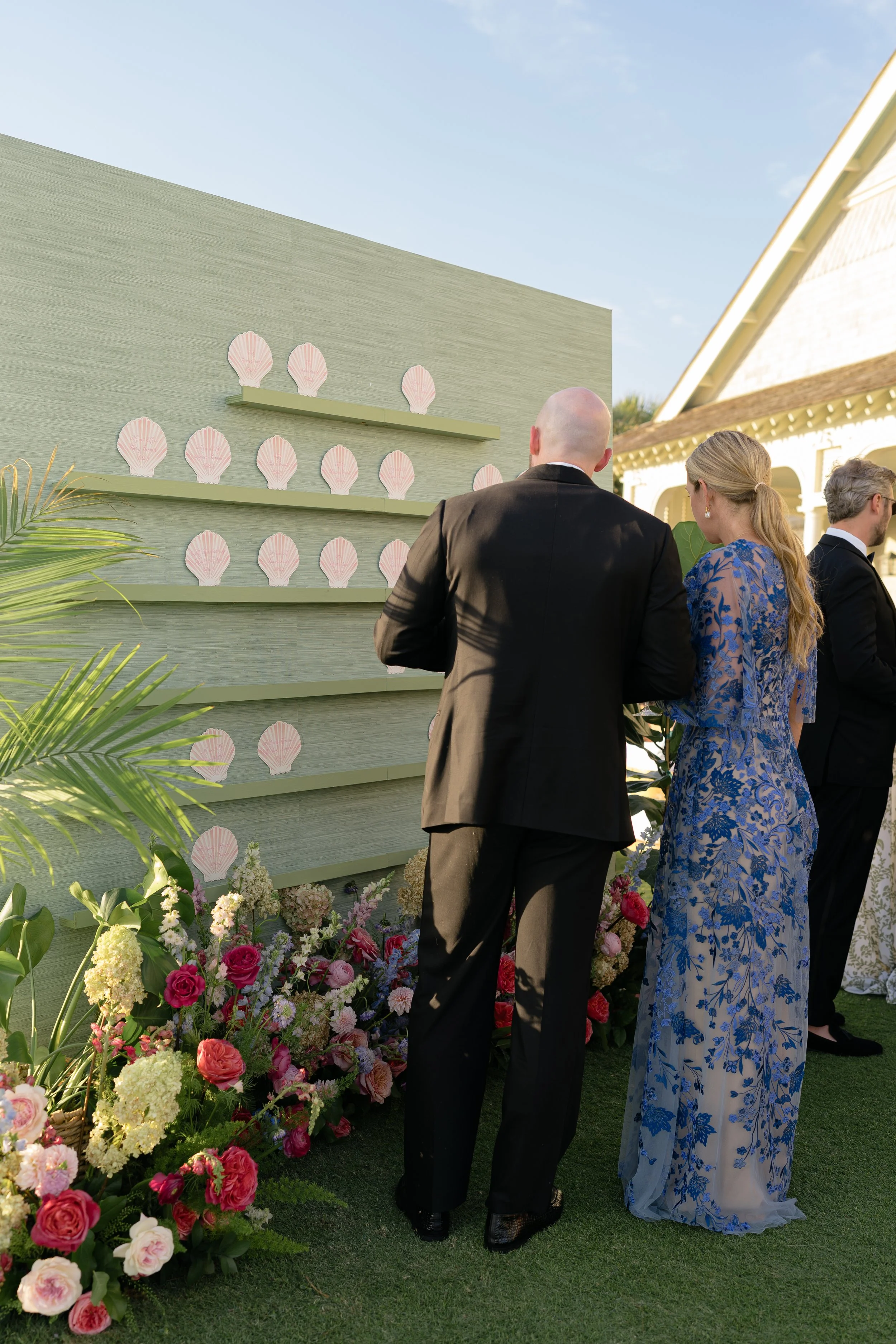Wedding guests find their seats at a pale green escort-card wall decorated with white seashells and tropical plants.