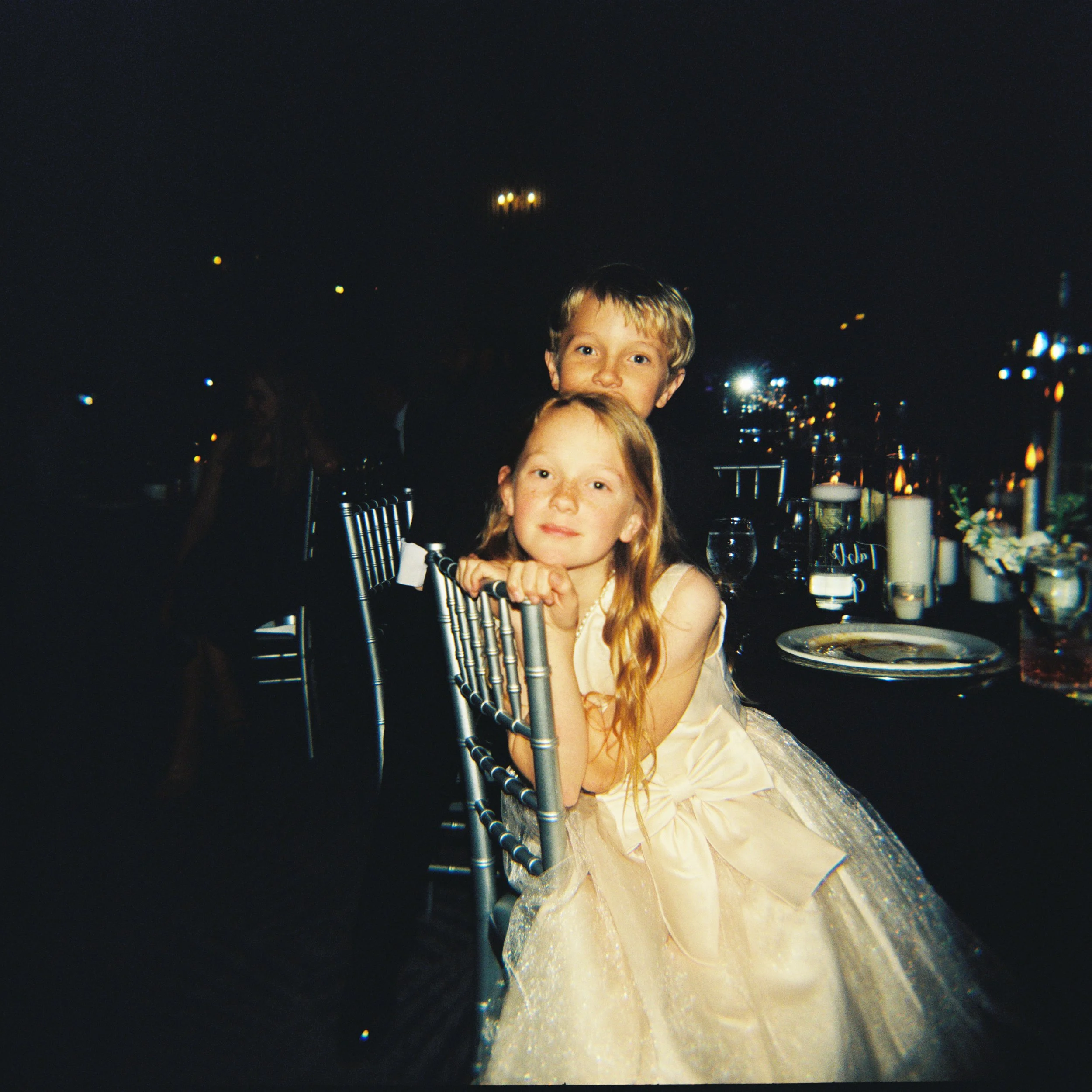 Young flower girl and her brother photographed during a wedding reception, illuminated by warm evening light and surrounded by the energy of the celebration.