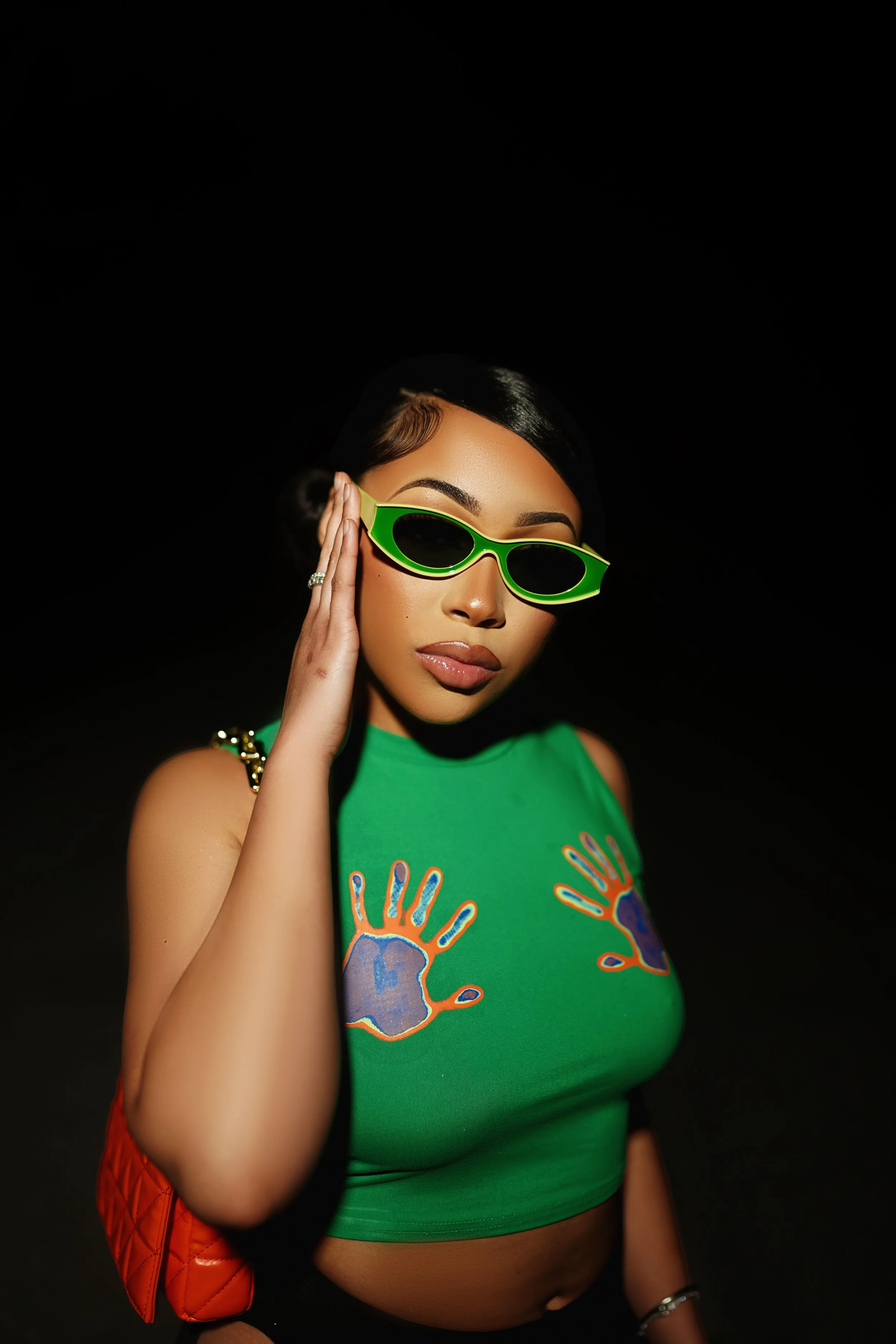 A woman wearing green sunglasses and a green sleeveless top with colorful handprints on it, posing against a black background.
