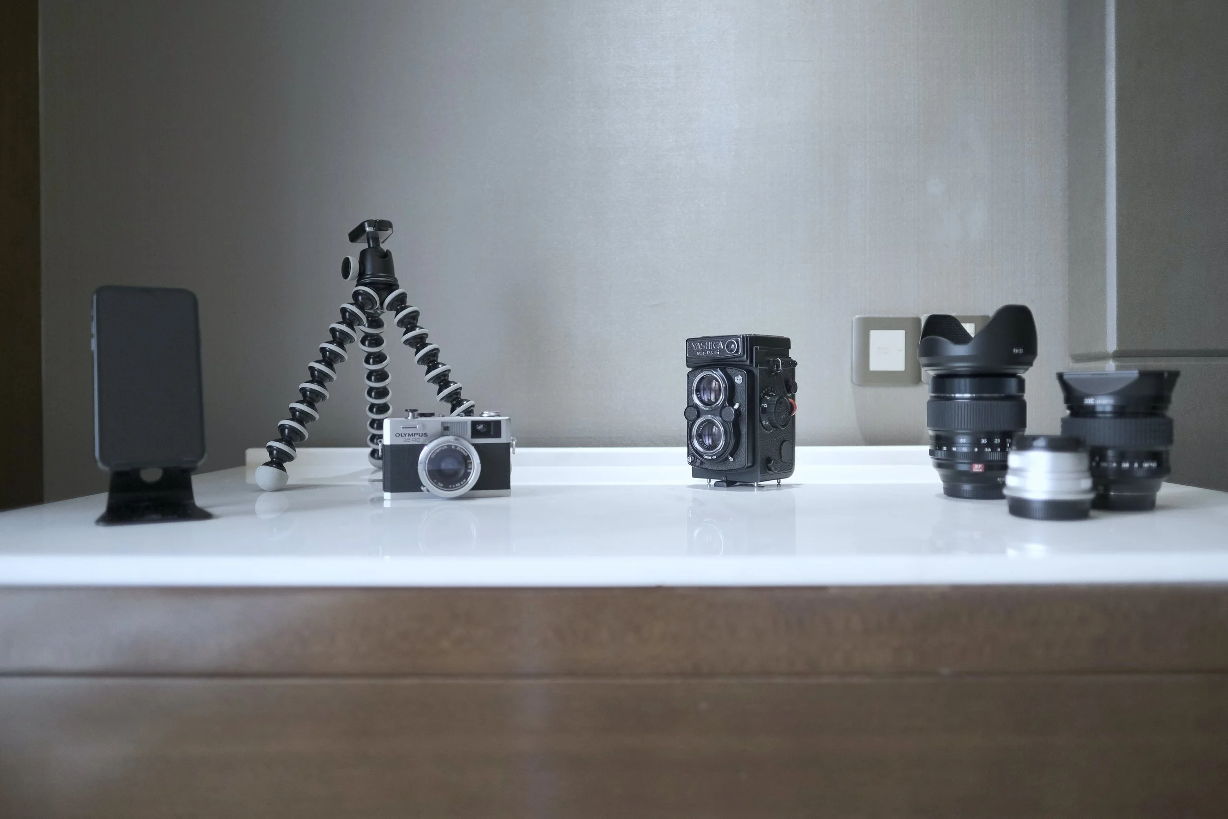 Collection of cameras and camera equipment on a white surface, including a small compact camera, a vintage twin-lens camera, a camera lens with a lens hood, a halogen bulb, and a flexible tripod.