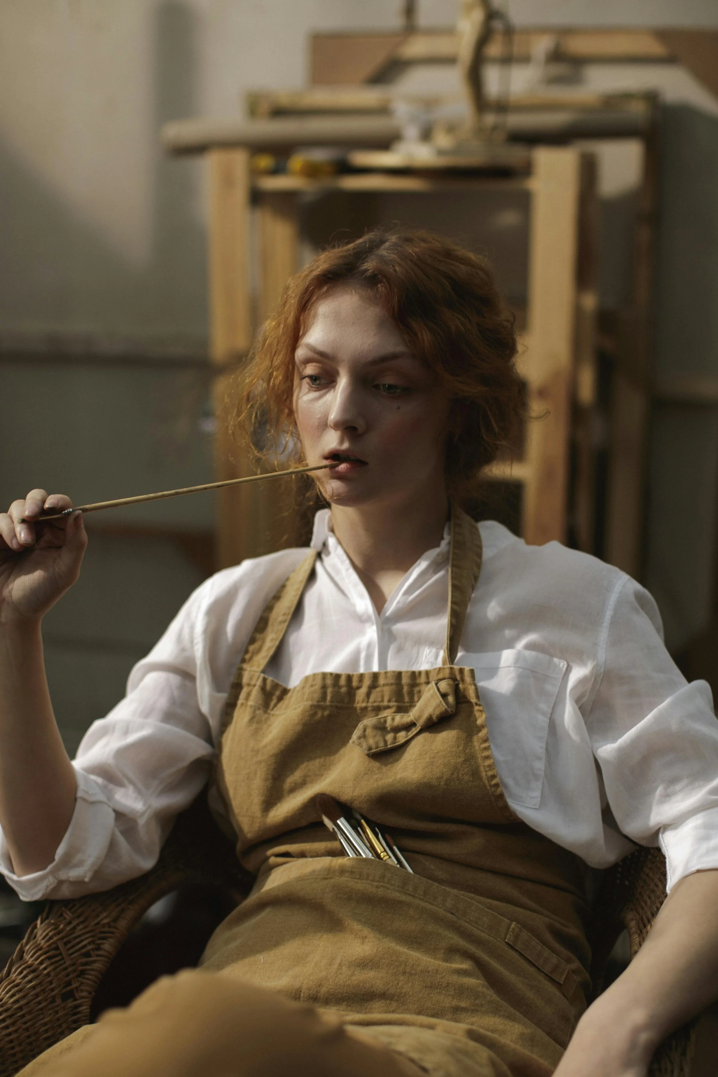A woman with short, curly red hair wearing a white shirt and an apron, holding a long stick in her mouth, sitting in a workshop with wooden structures in the background.