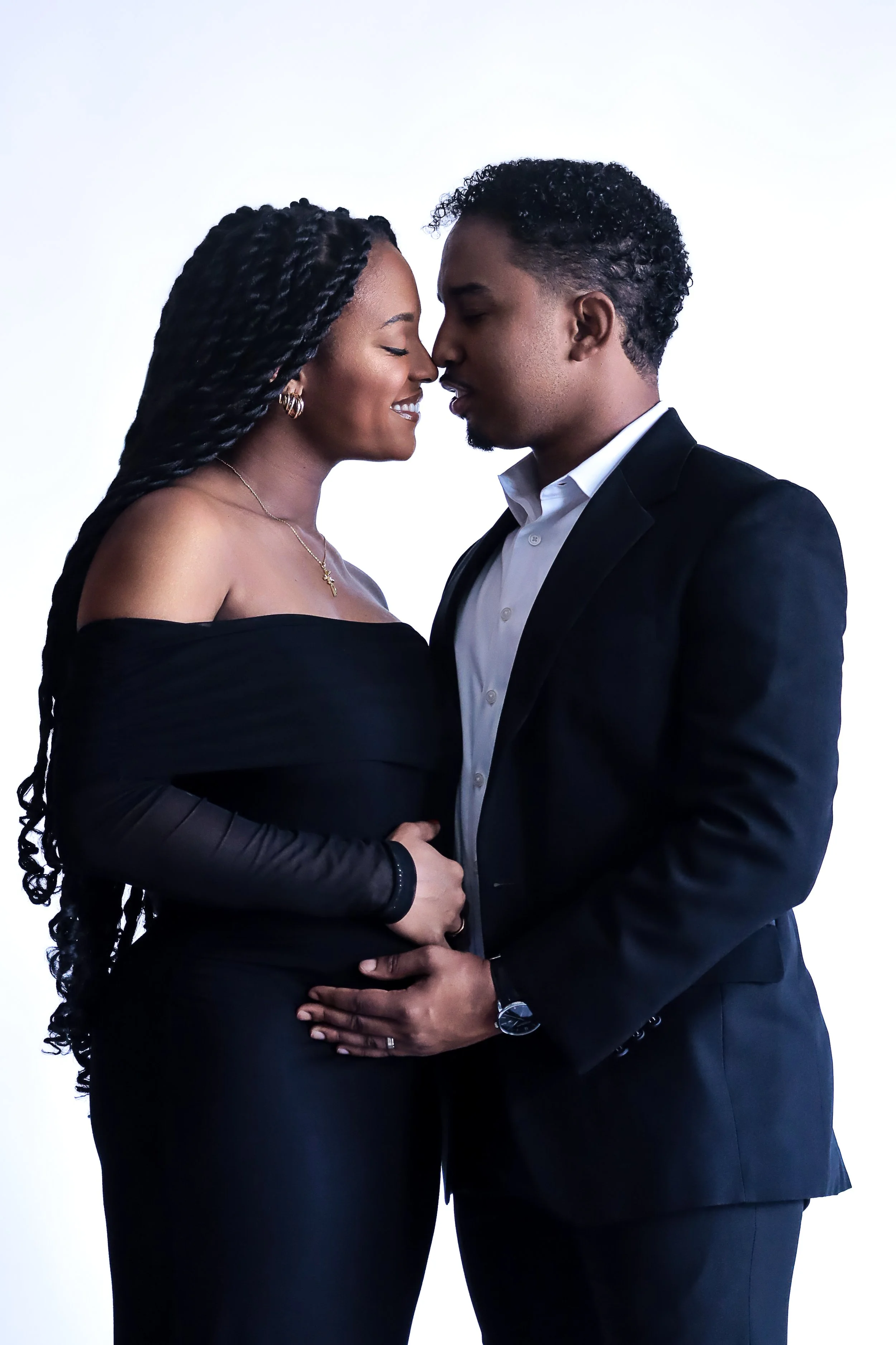 A man and woman in formal attire holding each other close, with foreheads touching, against a plain white background.