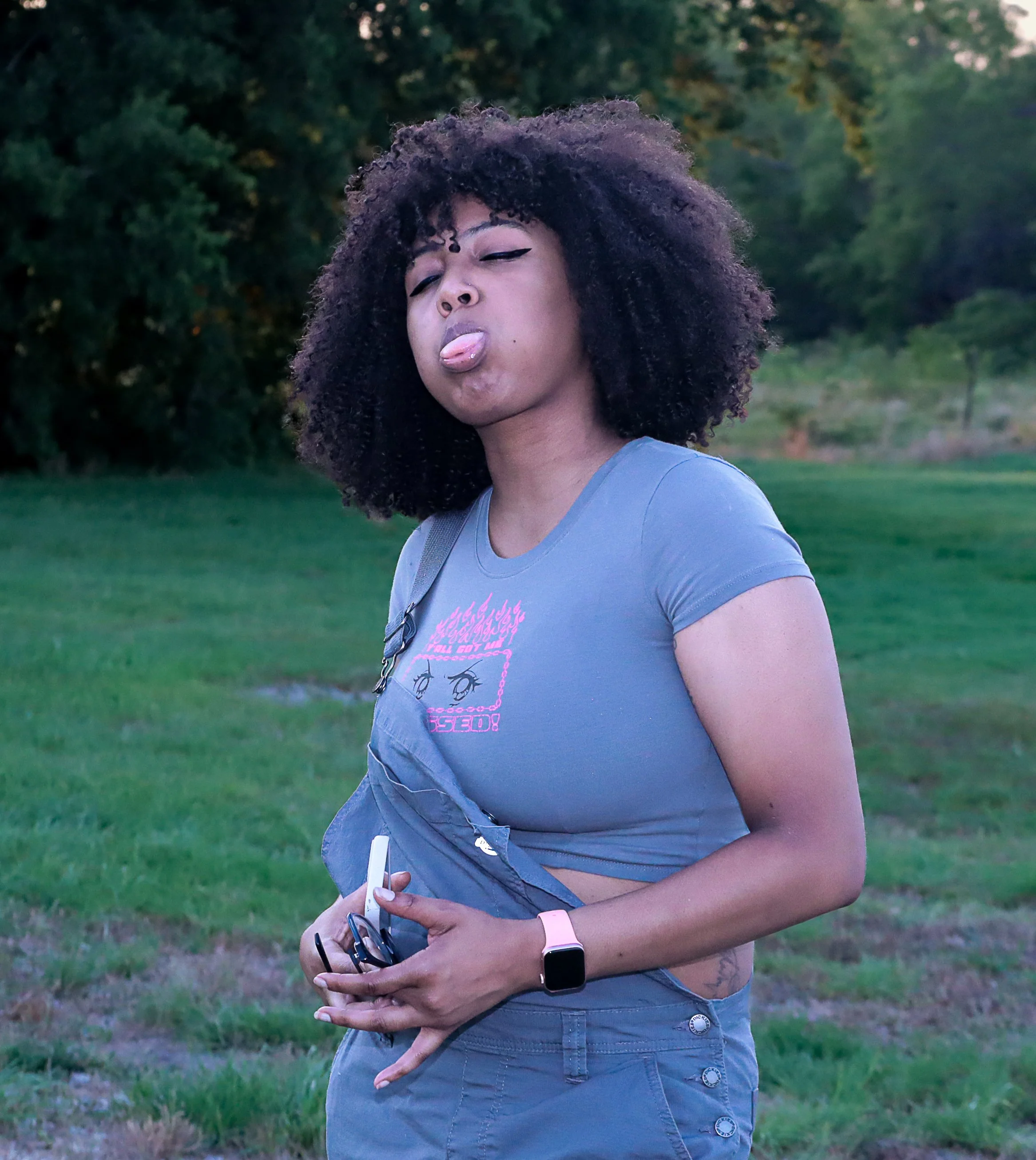 Young woman with curly hair in a park making a silly face by sticking out her tongue and closing her eyes.