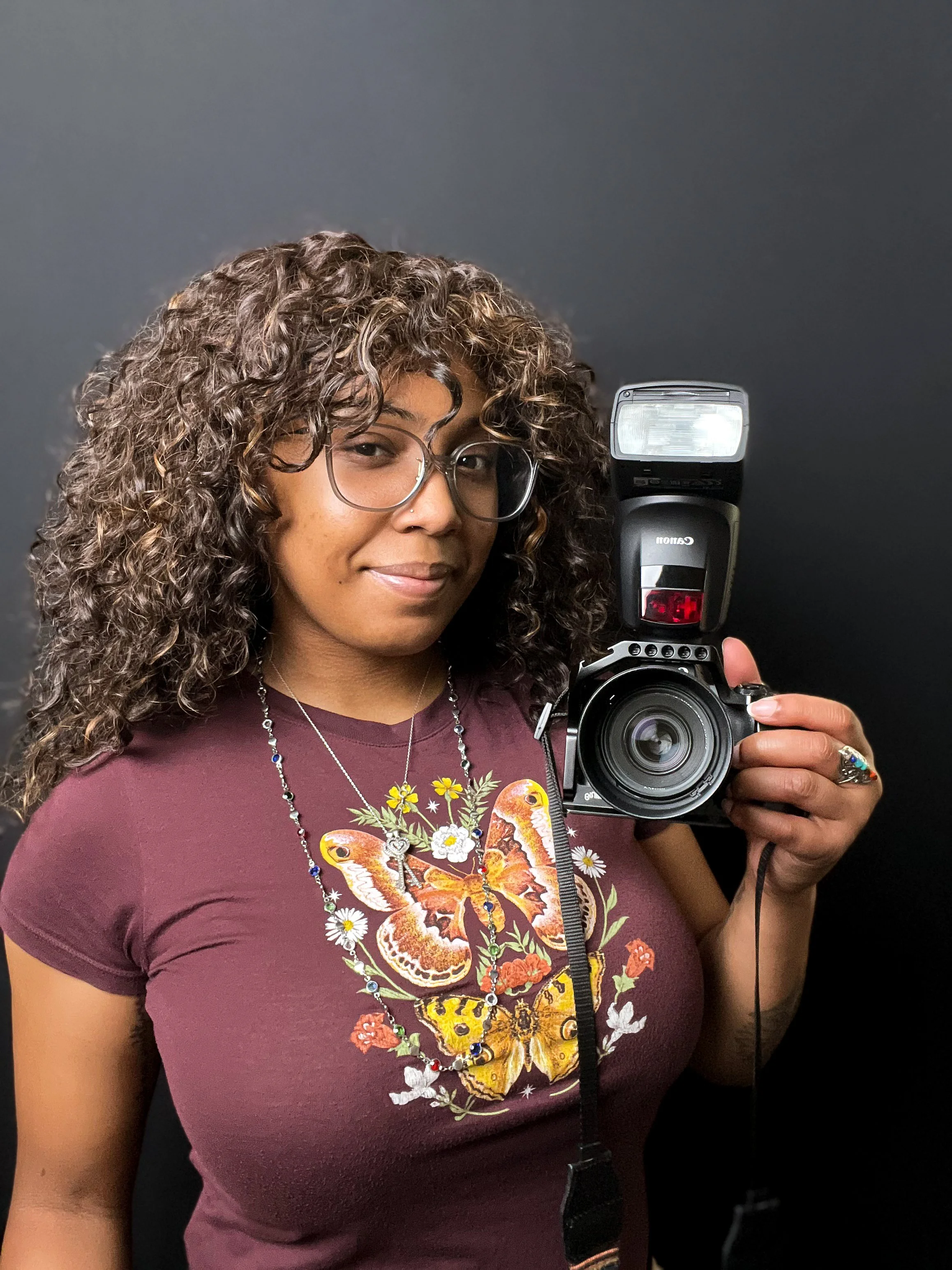 Headshot of photographer Jordan Fichter with curly hair and glasses, wearing a floral butterfly t-shirt while holding a camera with an external flash.