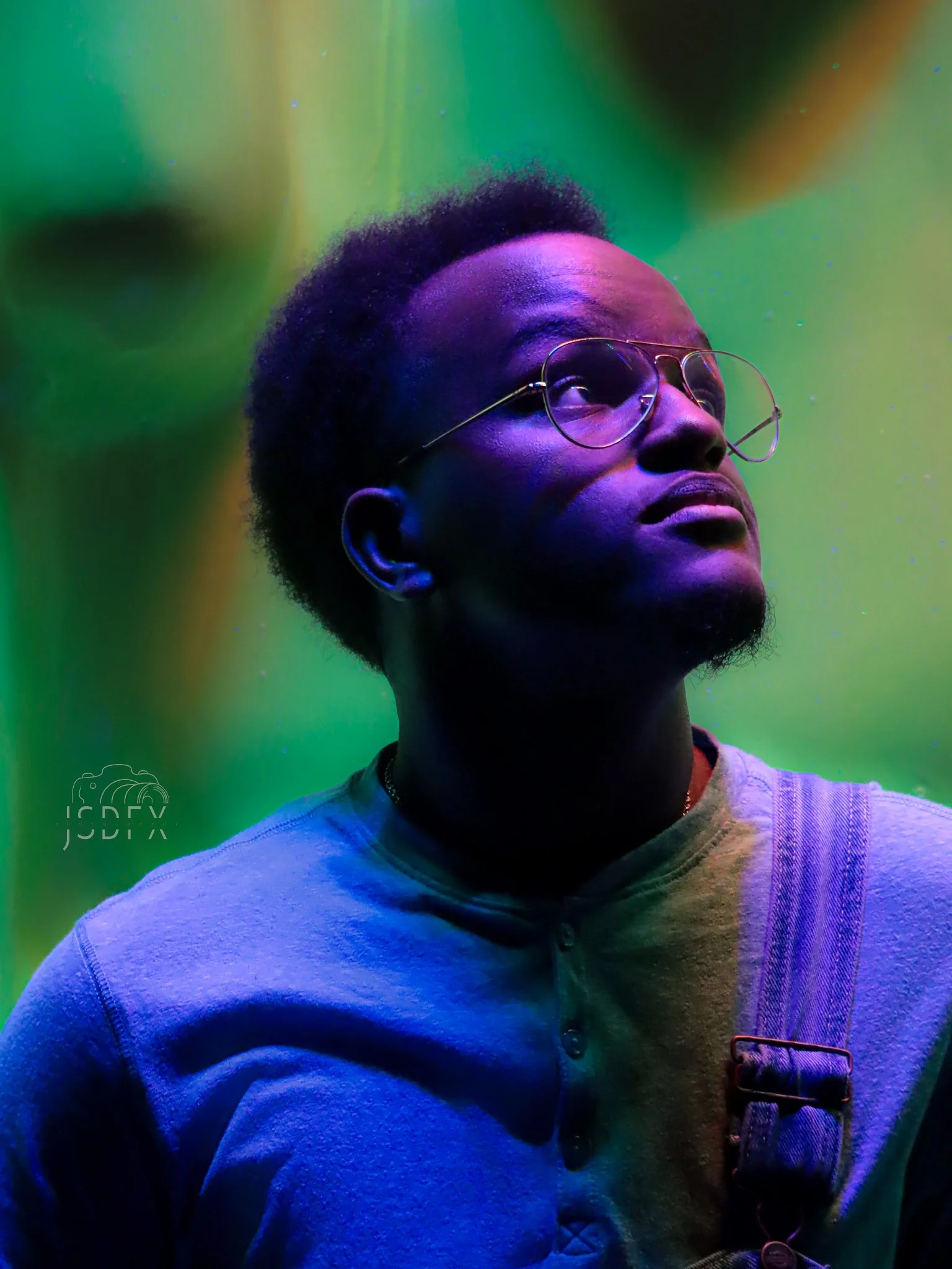 A person with glasses and short curly hair in a colorful, vibrant setting with green and purple lighting, looking upward.