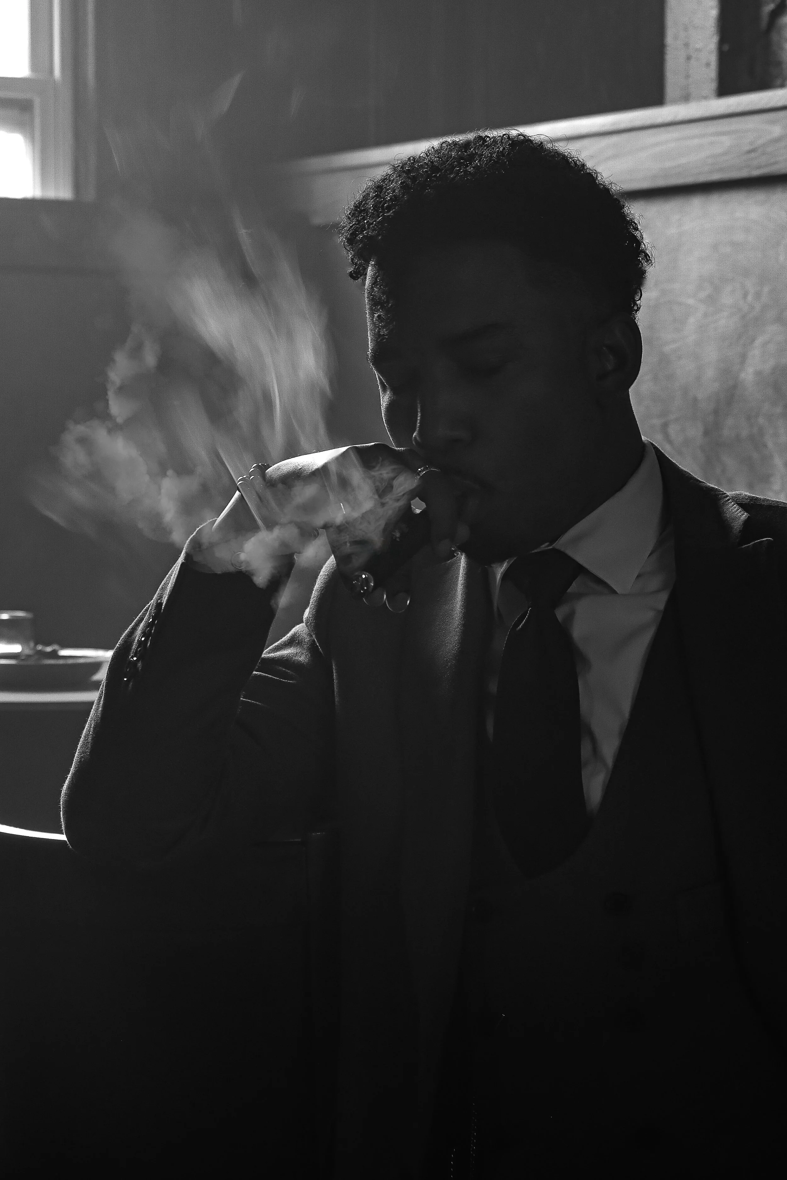 A man dressed in a suit and tie, smoking a cigar in a dimly lit room, black and white photo.