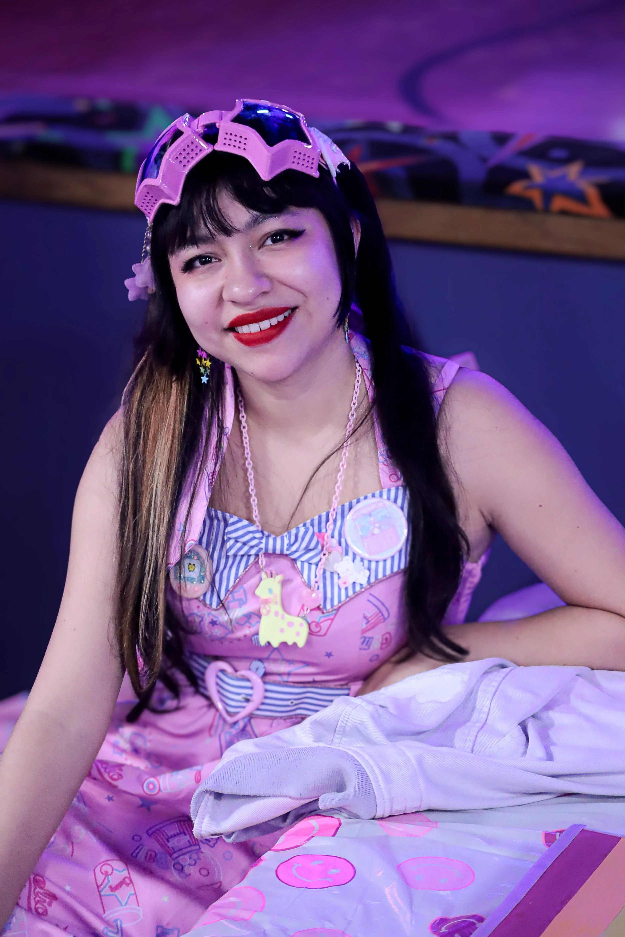 A woman with dark hair and bangs smiling, wearing pink roller skate goggles on her head, a pink dress with pastel decorations including unicorns and hearts, and star-shaped earrings, sitting on a bed with pink and white bedding in a room with purple and blue lighting.