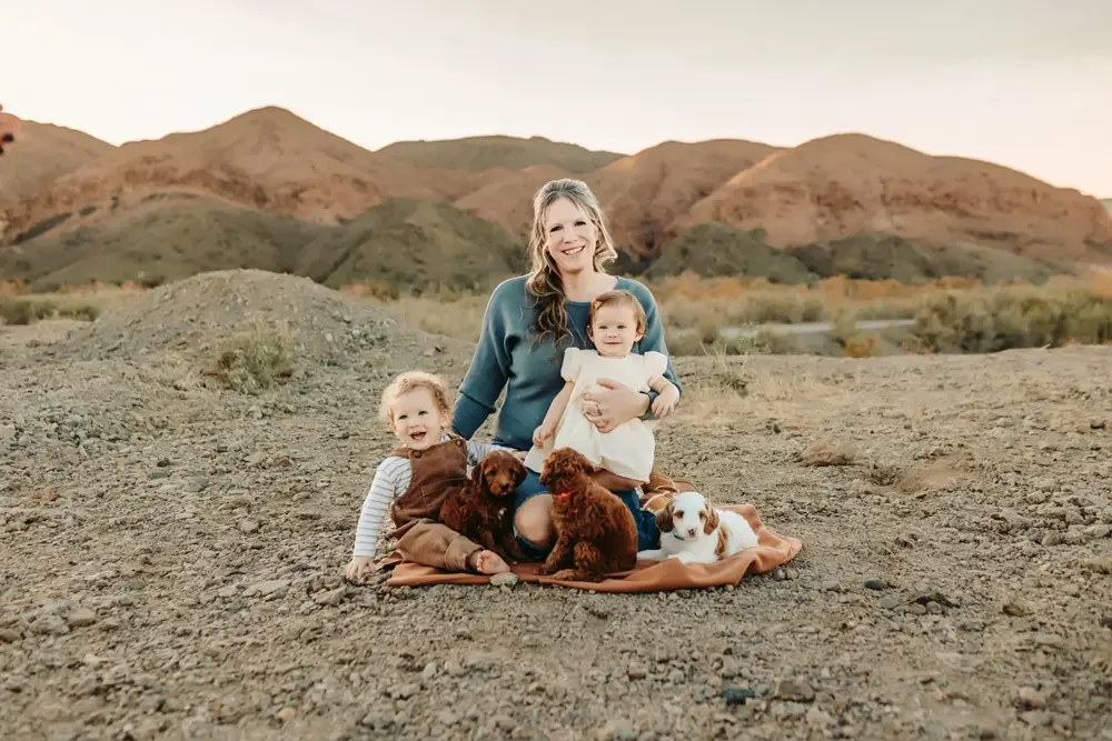 A woman with two young children and four puppies sitting on a blanket in a rocky outdoor area with mountains in the background.