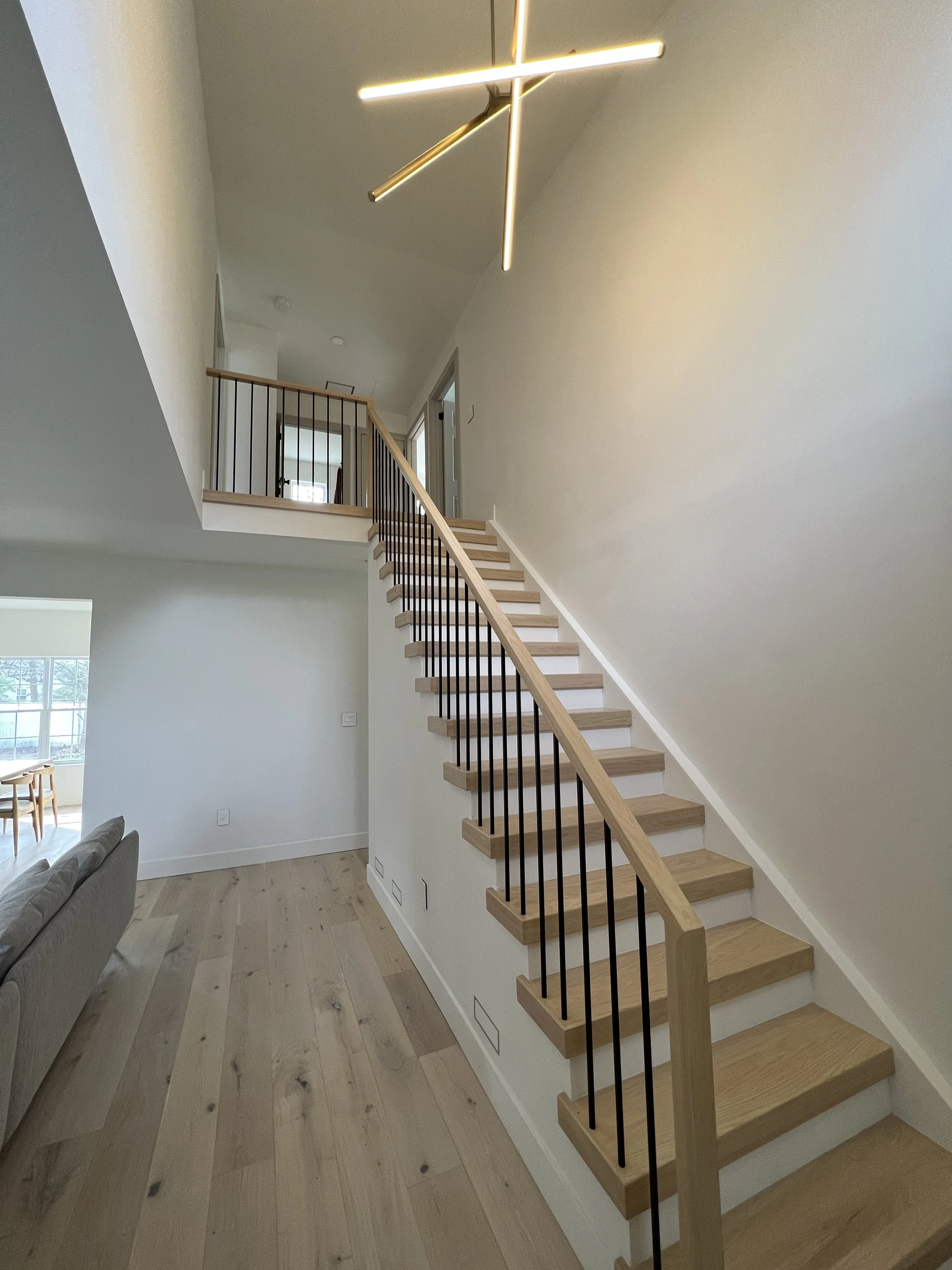 Interior view of a modern staircase with wooden steps, black metal balusters, and a wooden handrail, leading to an upper level with a balcony. Modern lighting fixture overhead.