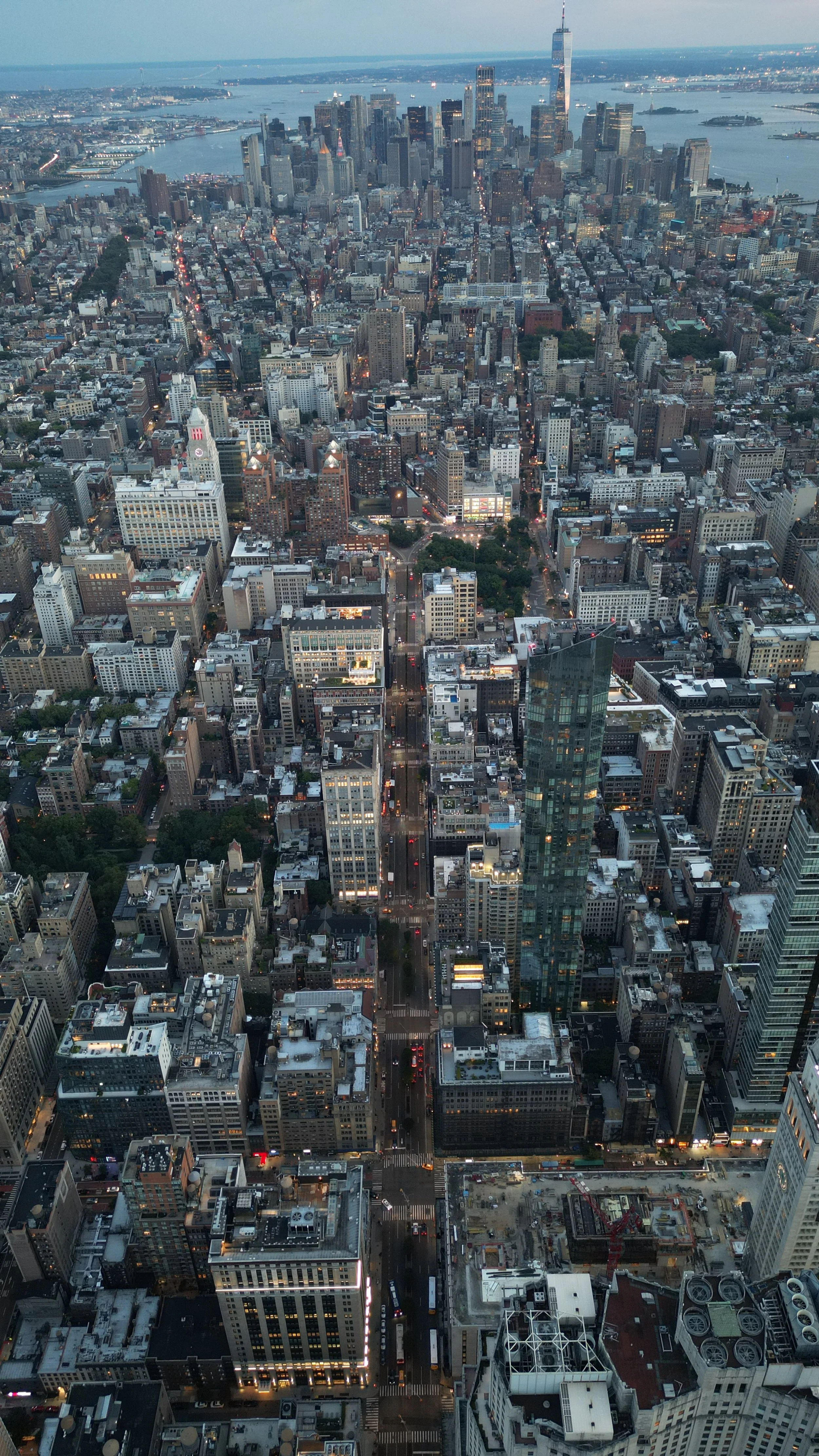 Aerial view of downtown New York City at dusk, showing numerous tall buildings, streets with traffic lights, and the Hudson River in the background.